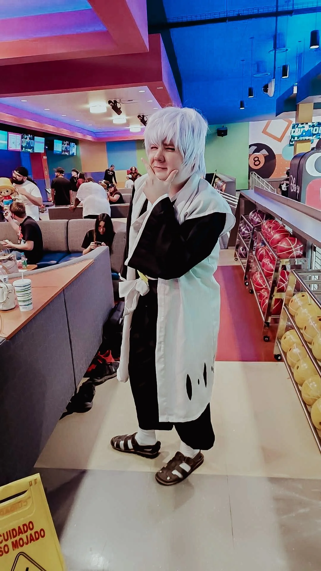 Person dressed in a black and white costume with white hair, posing playfully with their hand on their chin at an indoor entertainment venue with gaming and shopping area.