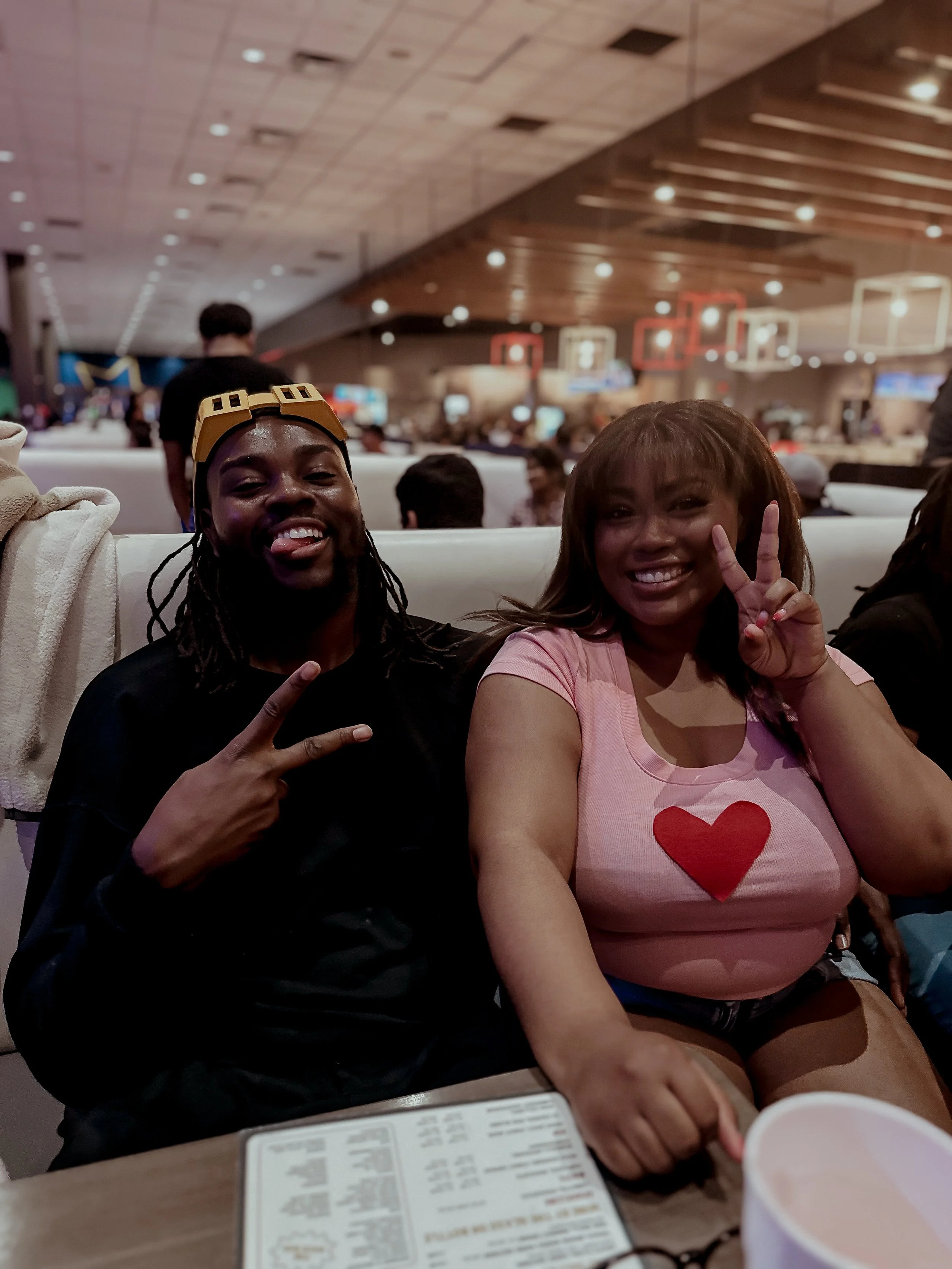 Two women sitting in a busy indoor setting, smiling and making peace signs with their hands, with a crowded background and ambient lighting.