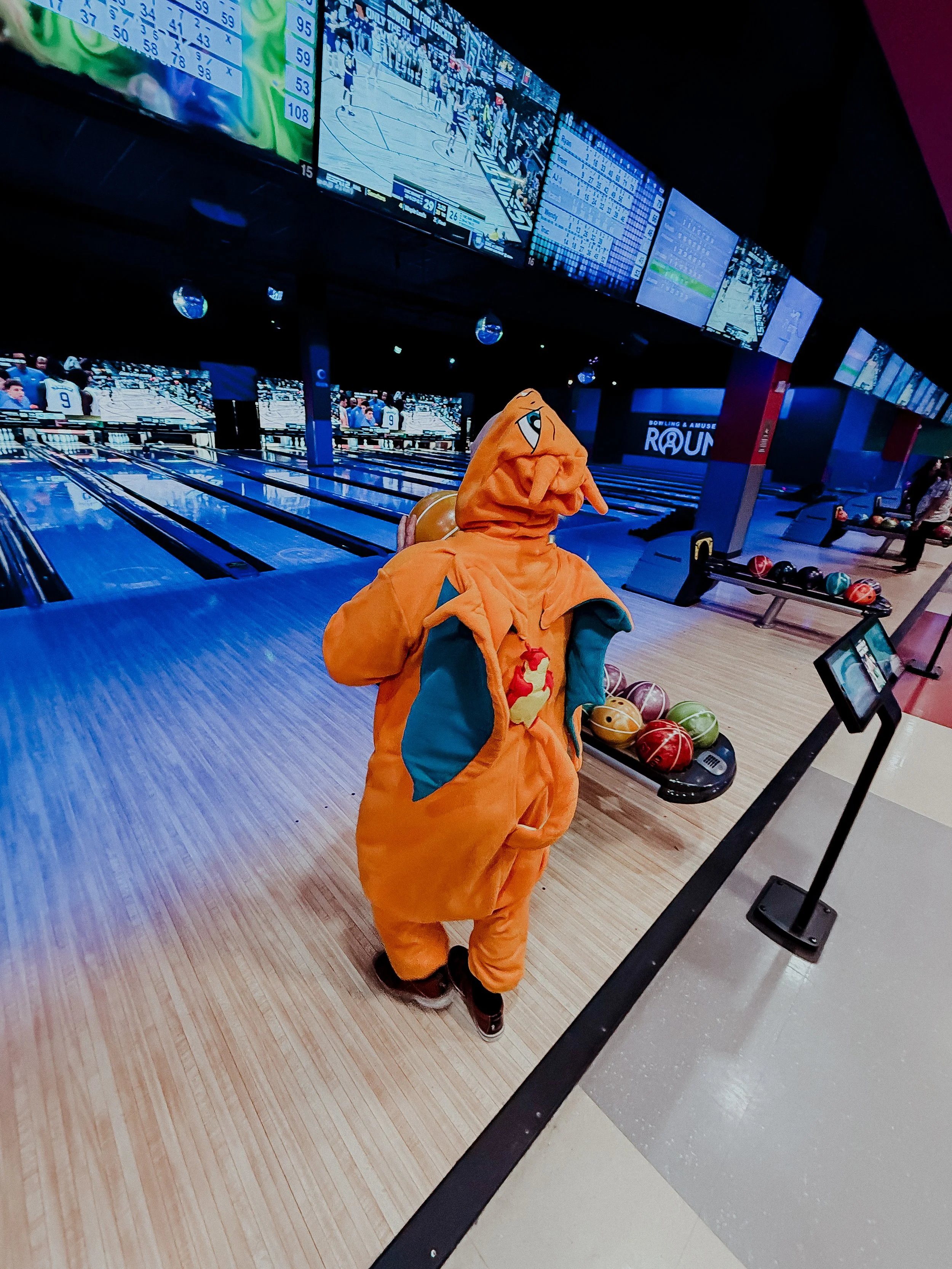 Person dressed in a large orange elephant costume standing on a bowling alley lane, holding the costume open to reveal a stuffed animal inside. Multiple bowling balls on a rack and electronic screens displaying scores and games are visible in the bac