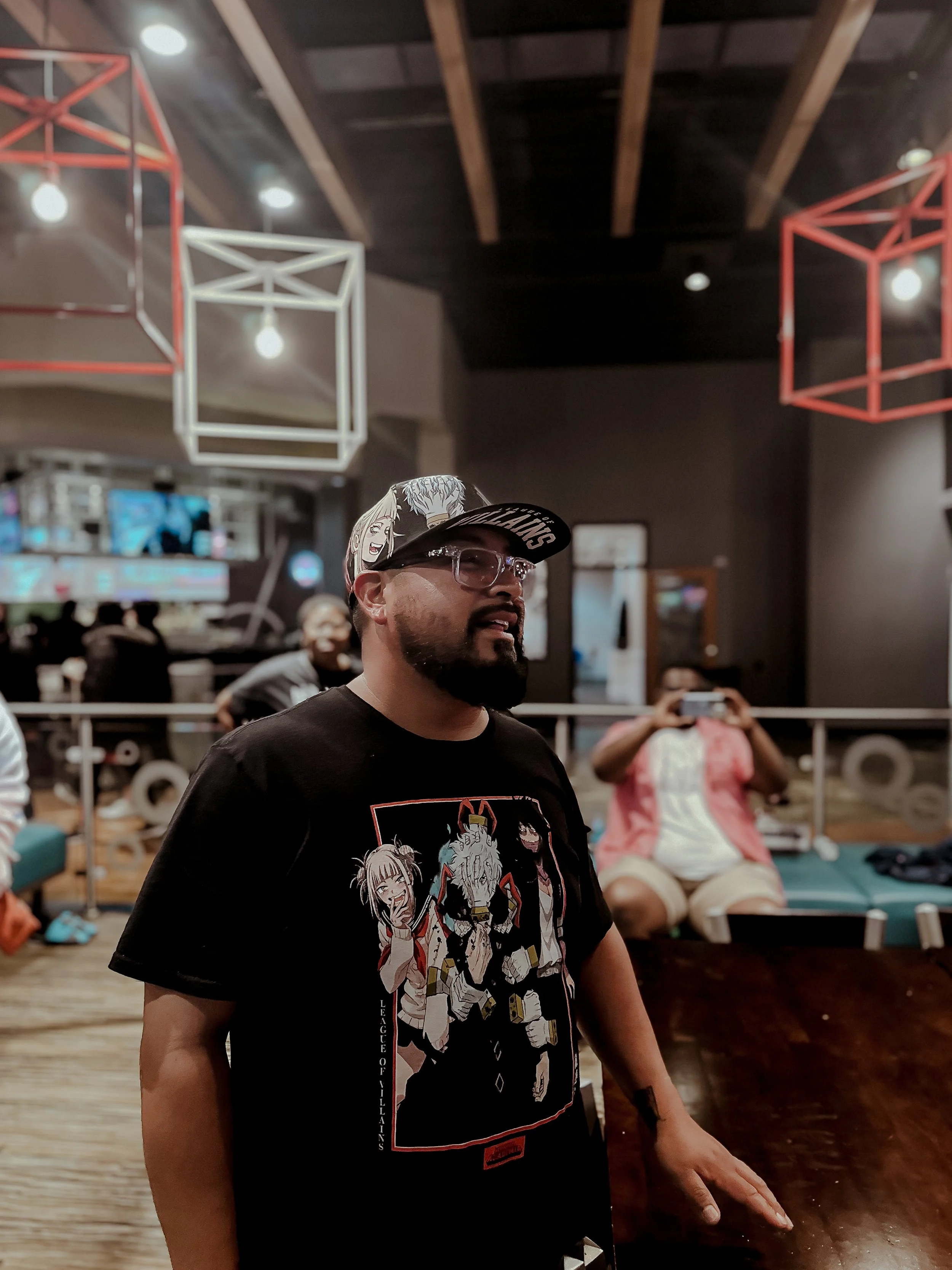 A man with glasses and a beard wearing a black graphic t-shirt and a black cap with white lettering, standing indoors at a social event or gathering. In the background, a person is taking a photo, and there are modern light fixtures hanging from the 
