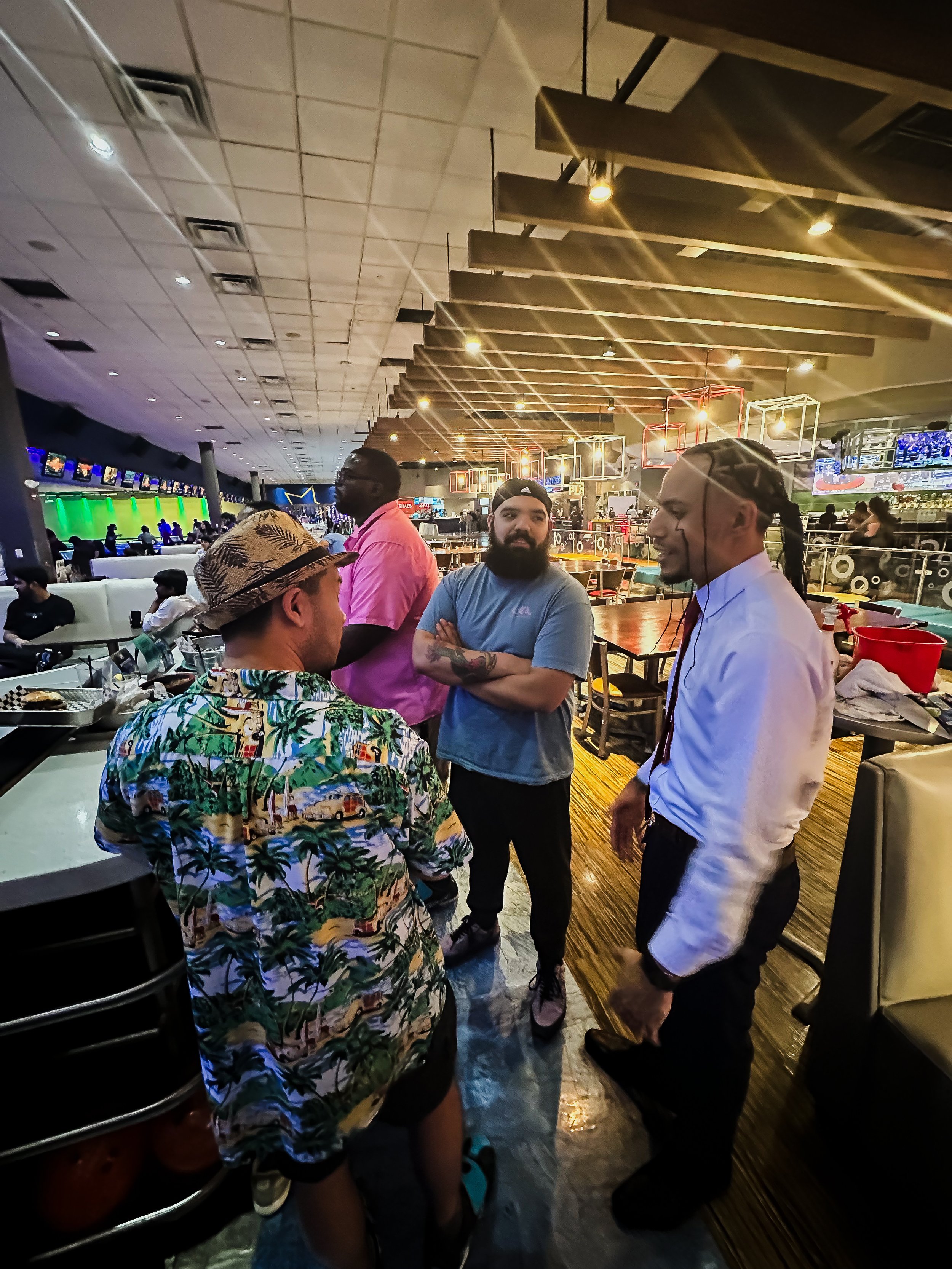 Four men engaged in conversation inside a sports bar with multiple screens showing sports games, illuminated in vibrant colors, and a busy restaurant atmosphere.