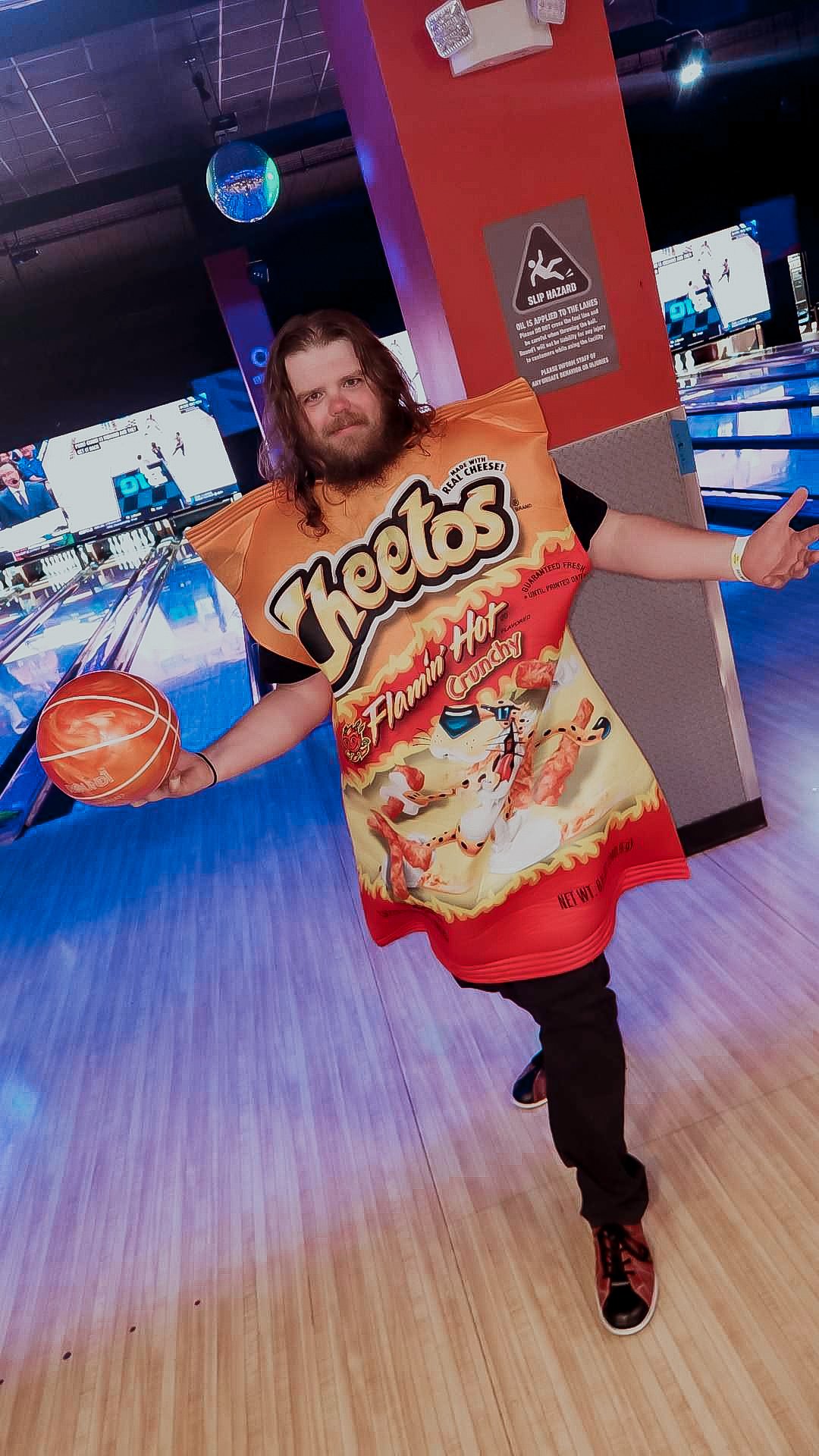 A man dressed in a T-shirt resembling a Bag of Nacho Cheese Doritos holding a basketball at a bowling alley.