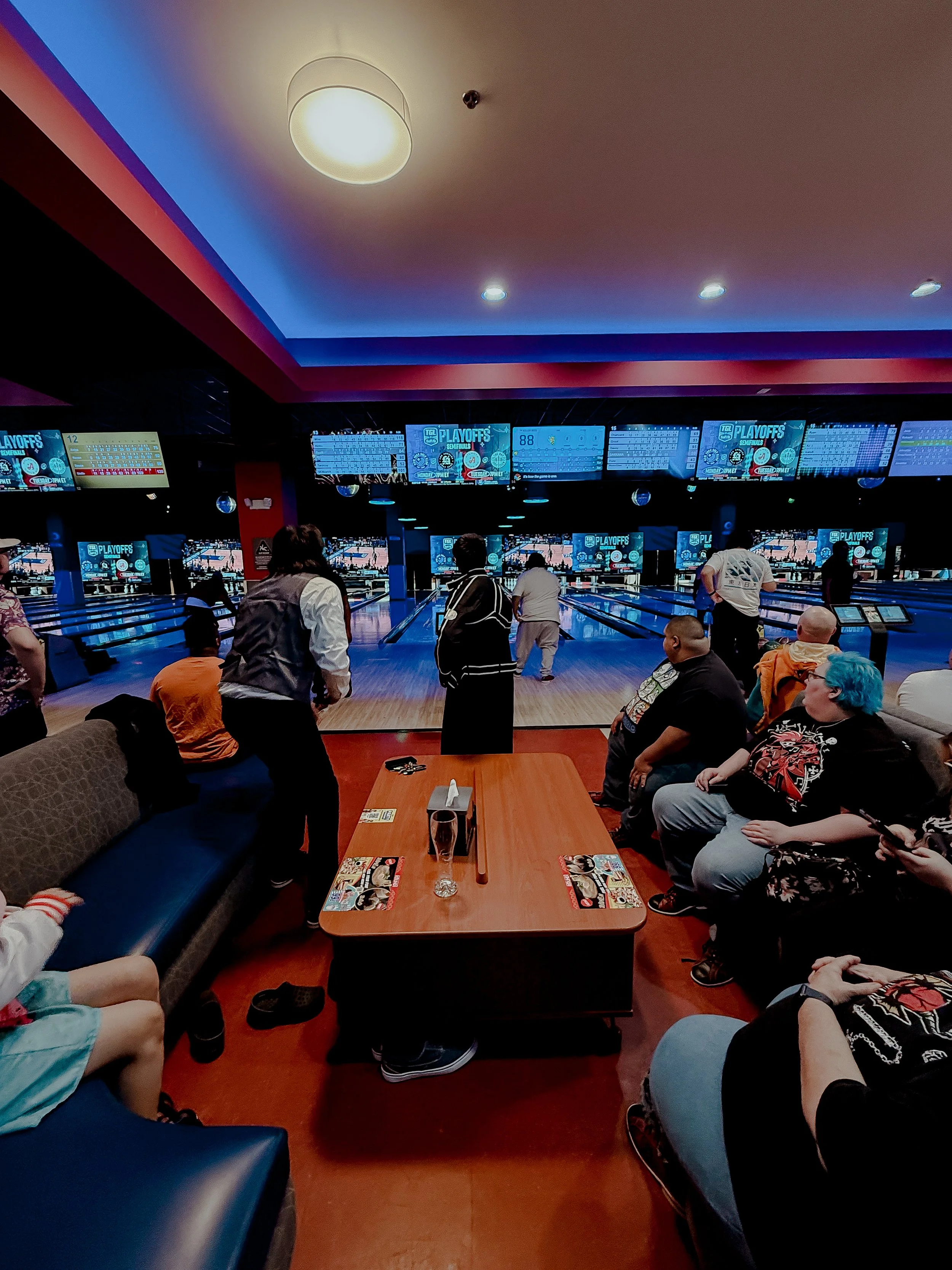 People sitting and watching a bowling alley with multiple screens displaying scores and league information.