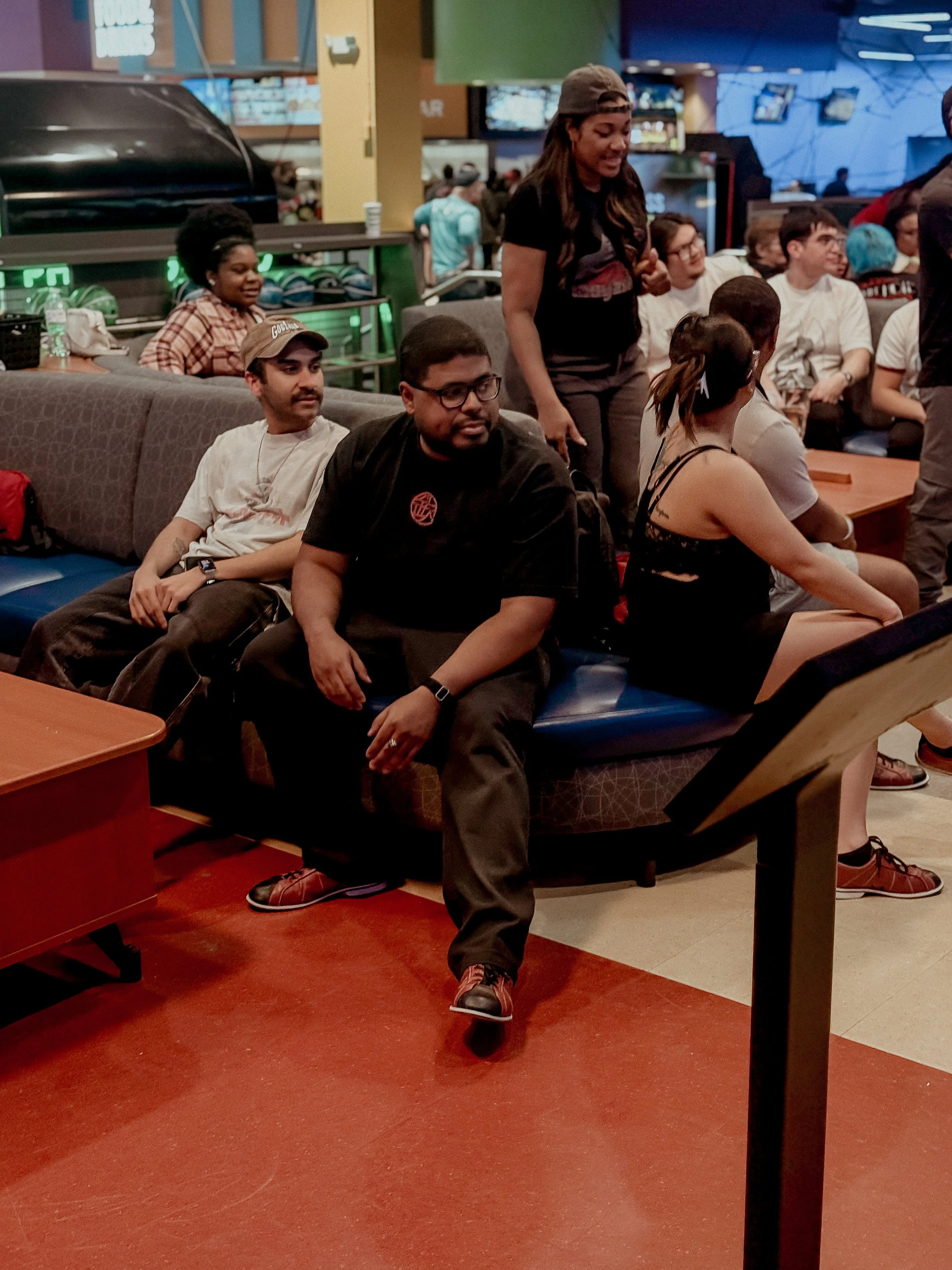 A group of people seated and standing at a bowling alley, with some engaged in conversation and others waiting for their turn to bowl.