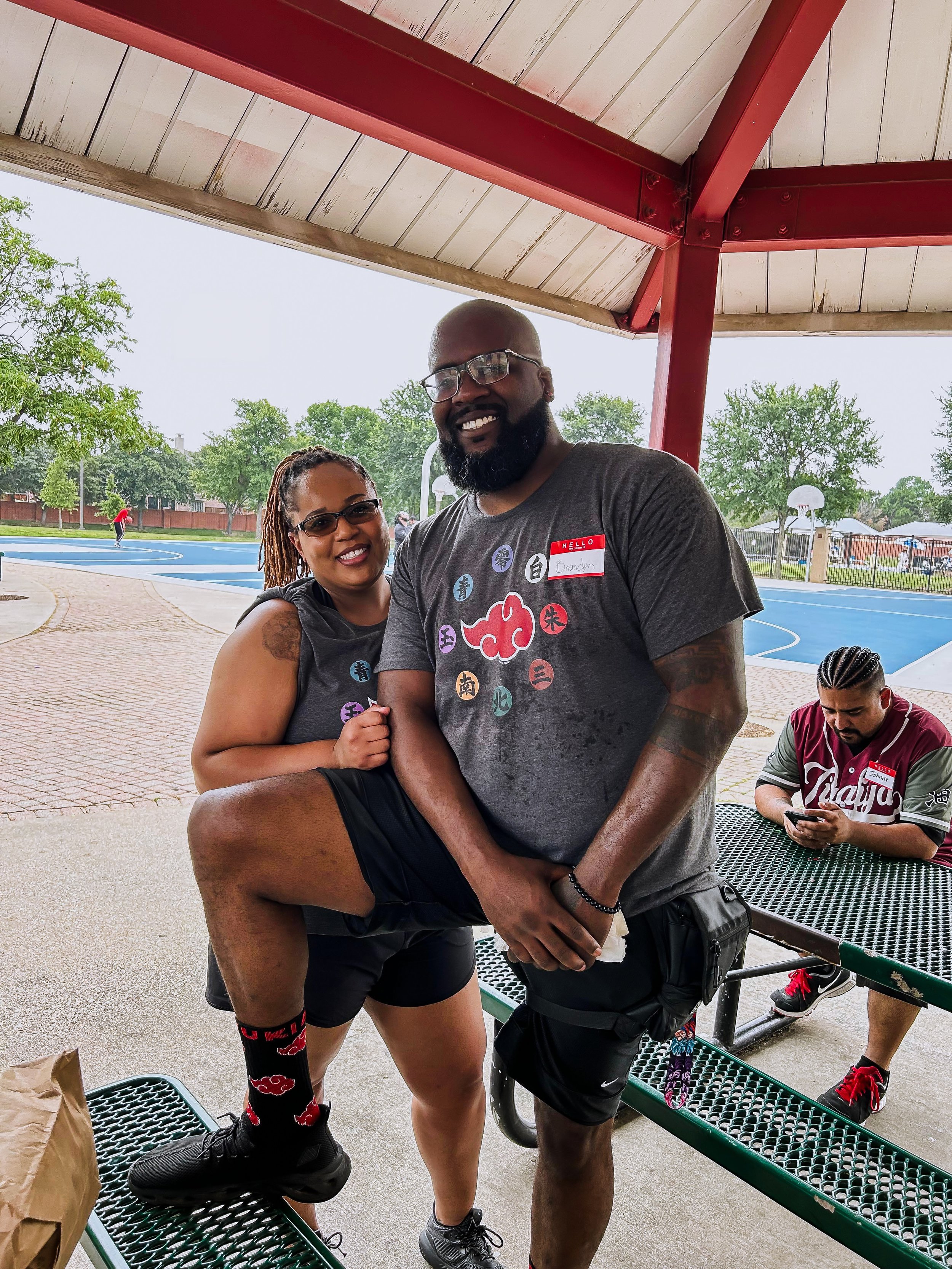 Two people smiling at a park under a pavilion, one with their leg raised and resting on a bench, the other standing next to them. In the background, a man is sitting at a picnic table looking at his phone, and a basketball court with a person playing