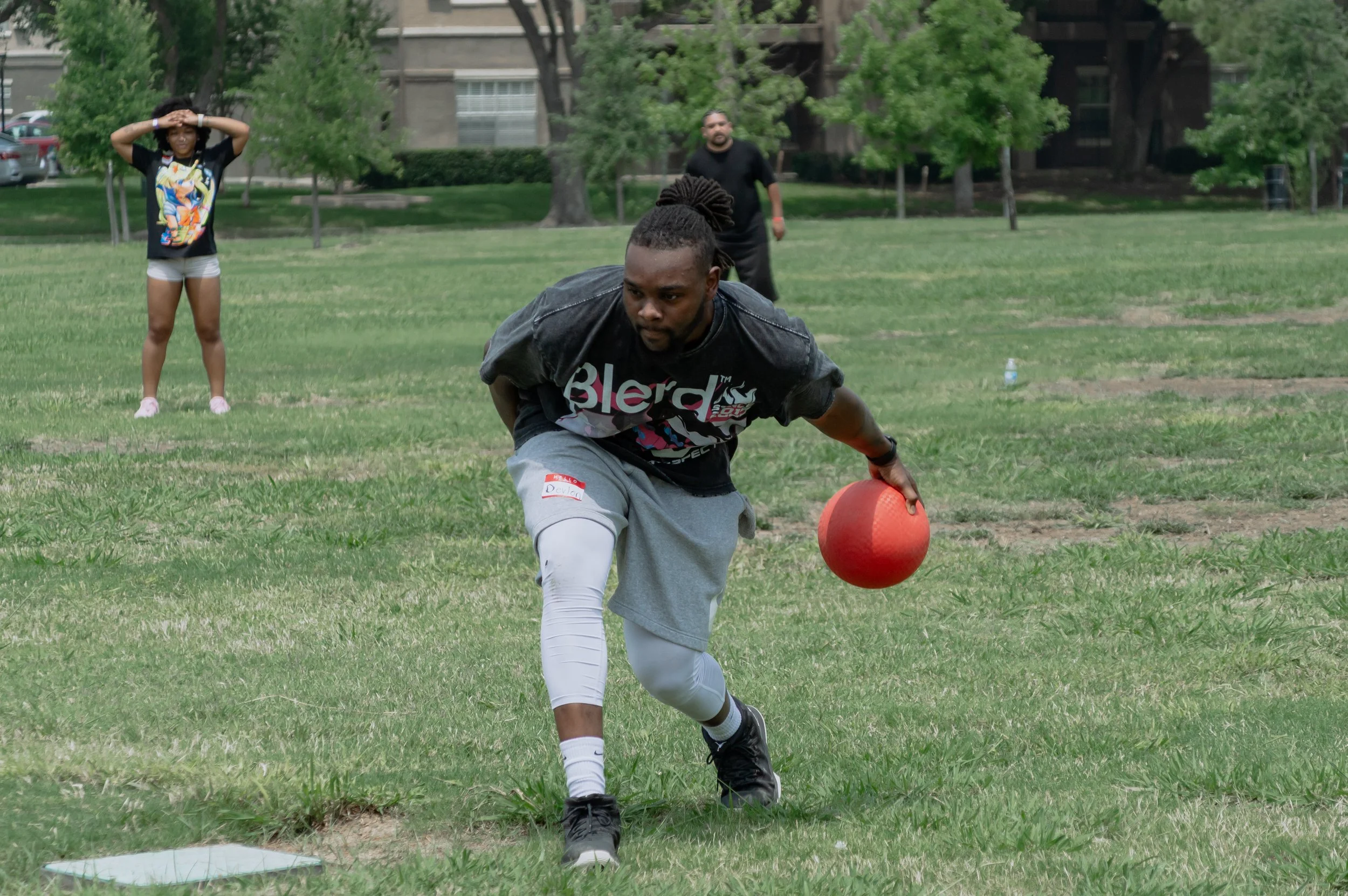 A football player in a black t-shirt and gray shorts is holding a red football while crouching on a grassy field. In the background, a woman in shorts and a graphic t-shirt stands with her hands on her head, and a man in a black shirt stands watching