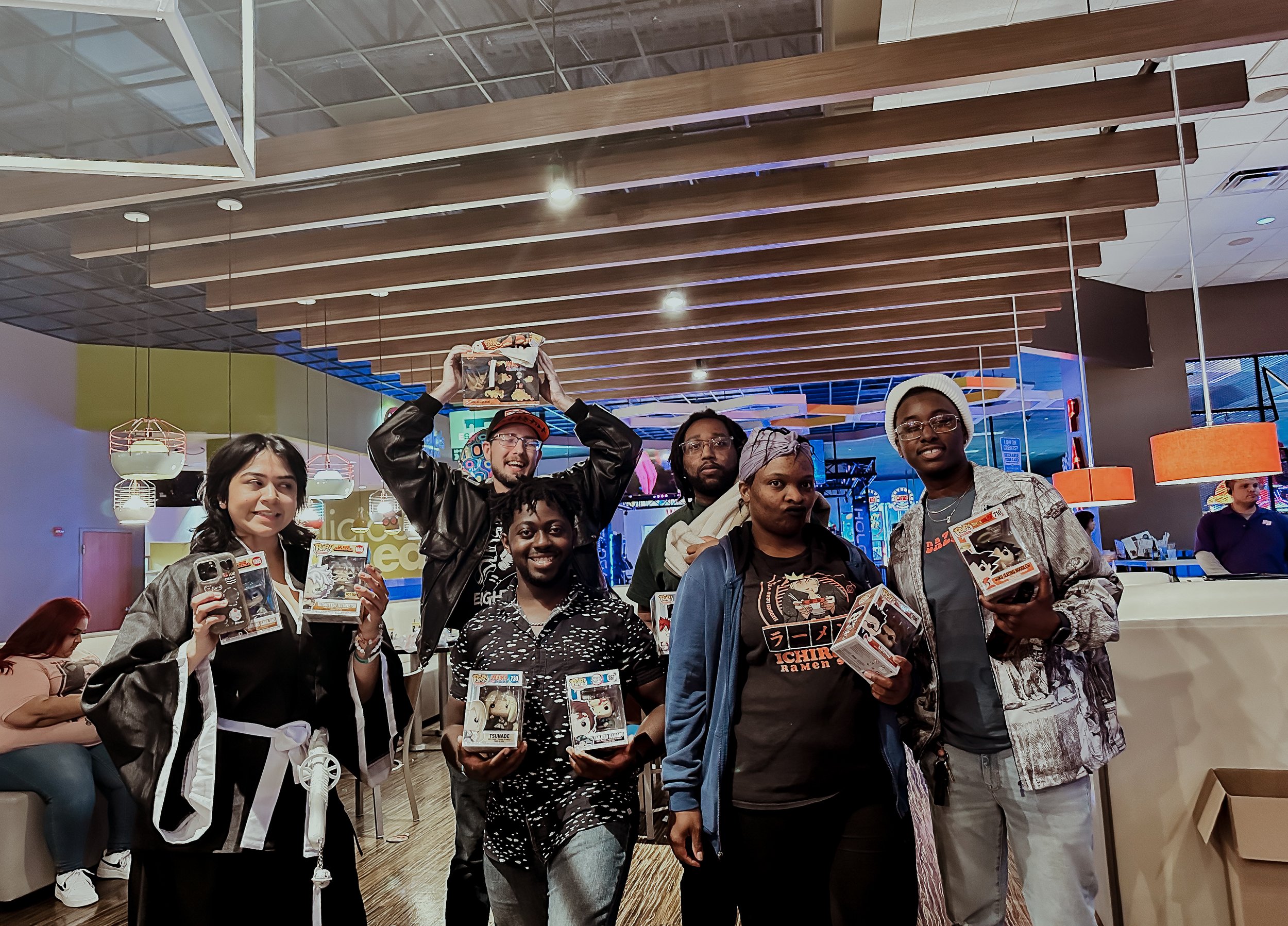 Group of six people smiling and holding Funko Pop figures at an indoor entertainment venue or arcade.