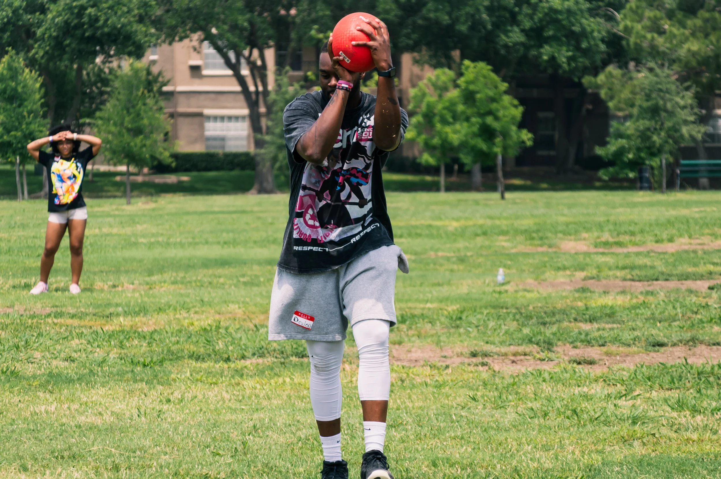 A young man in athletic clothes holding a red medicine ball above his head in a grassy park with trees and a building in the background. A young woman in casual sportswear stands in the background watching.