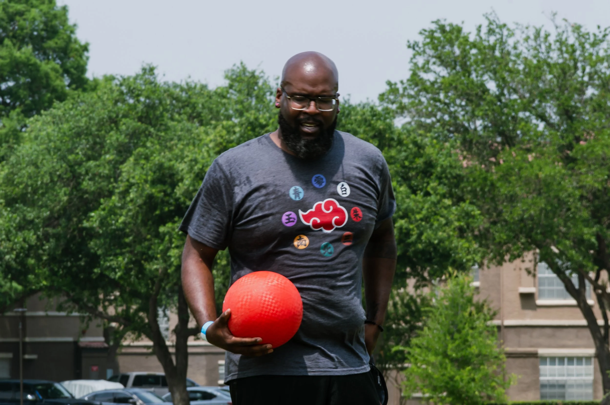 A man with glasses and a beard, wearing a dark gray t-shirt with colorful Japanese characters, holds a red exercise ball outdoors on a sunny day with green trees and a building in the background.