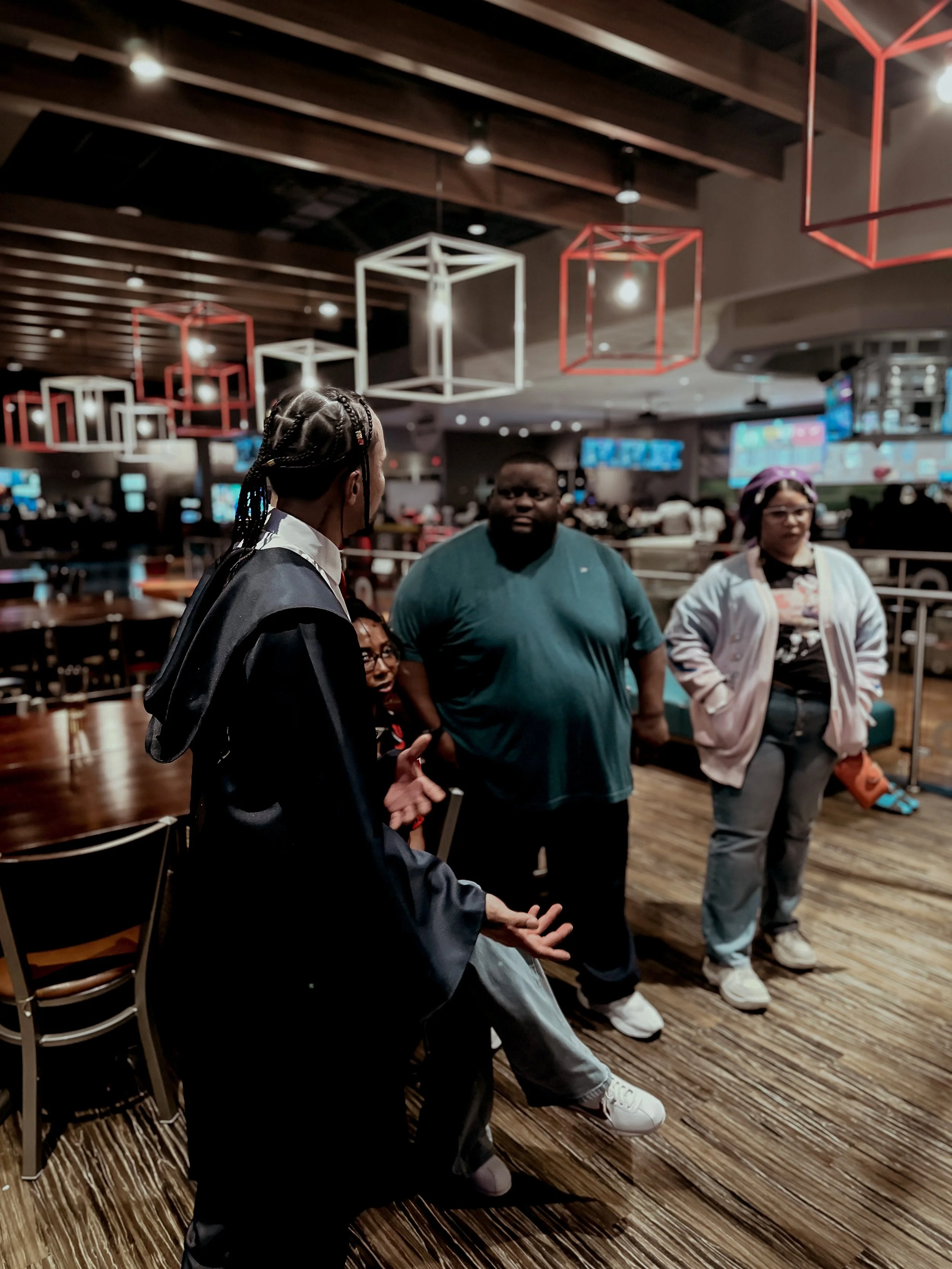 A woman in a graduation gown speaking to three people in a restaurant with wooden floors and modern lighting fixtures.