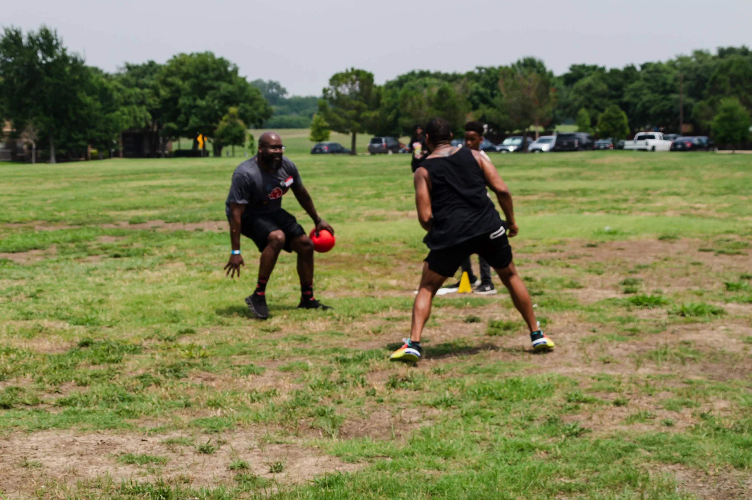 Two men playing outdoor football with a red ball on a grassy field in a park with trees and parked cars in the background.