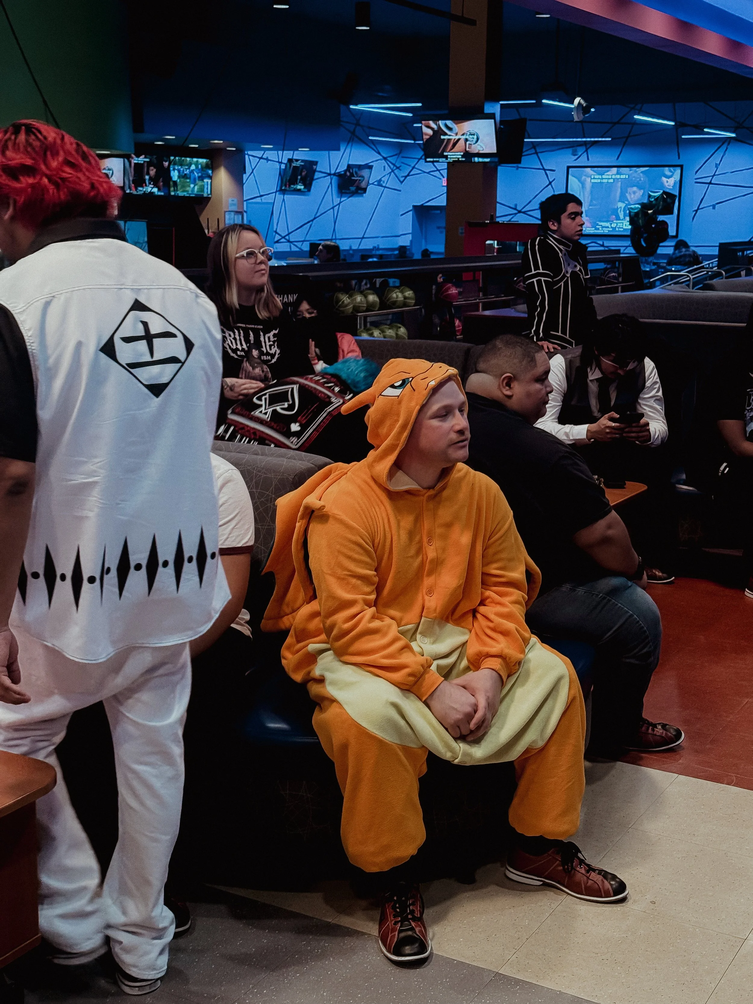Person dressed in a Charzard costume sitting on a bench in a bowling alley, surrounded by other people, some using smartphones, with televisions and neon lights in the background.