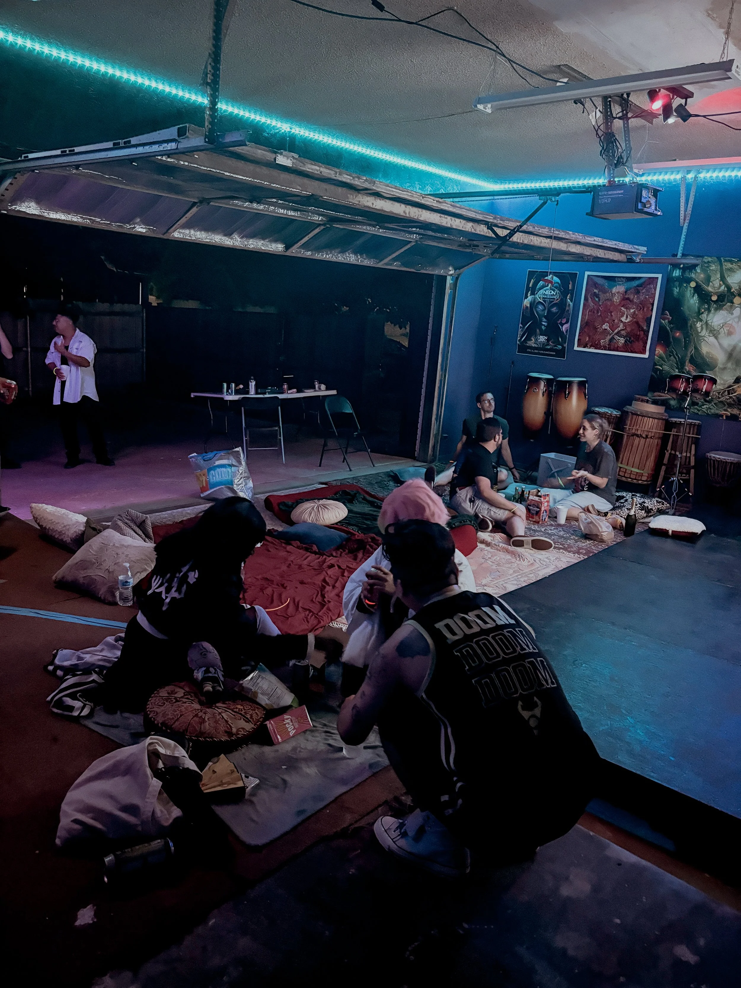 People sitting on blankets and cushions inside a dimly lit space with musical instruments, posters, and blue neon lighting.