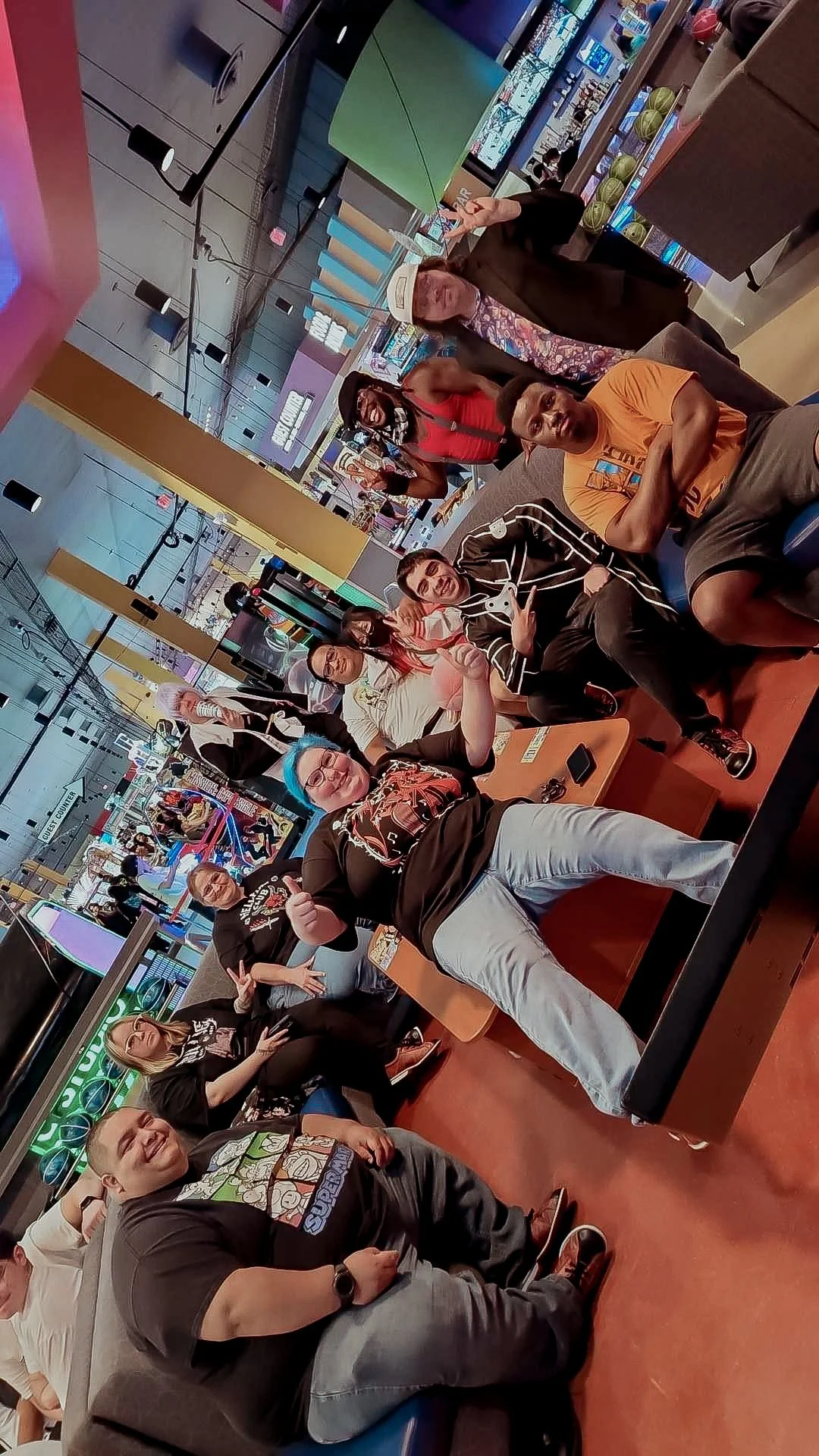 Group of people sitting and standing in a colorful arcade, with game machines and screens in the background, some making peace signs and smiling.