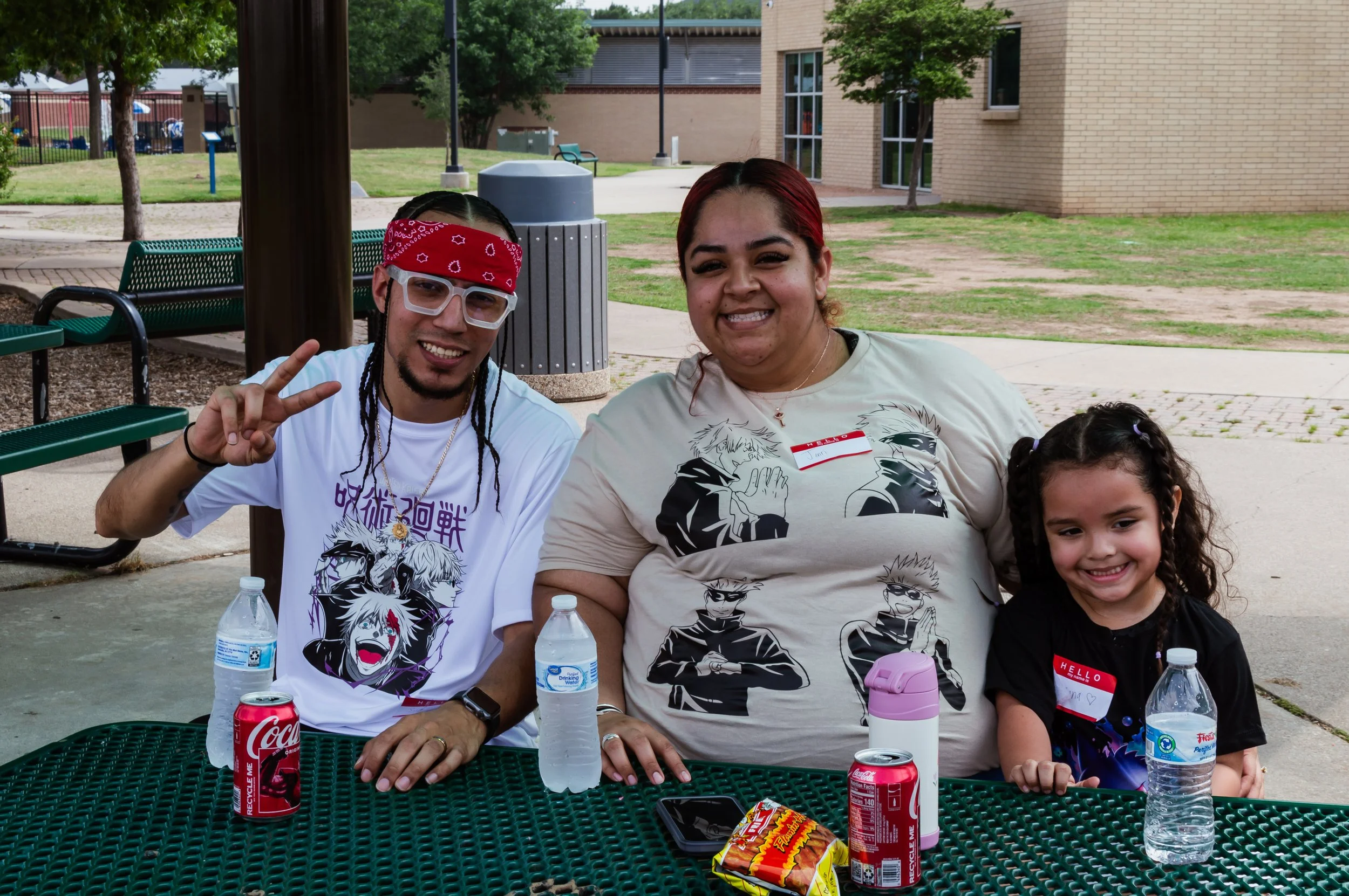 A group of three people, one young man, one woman, and a young girl, sitting at an outdoor table with drinks and snacks, smiling and posing for the photo in a park.
