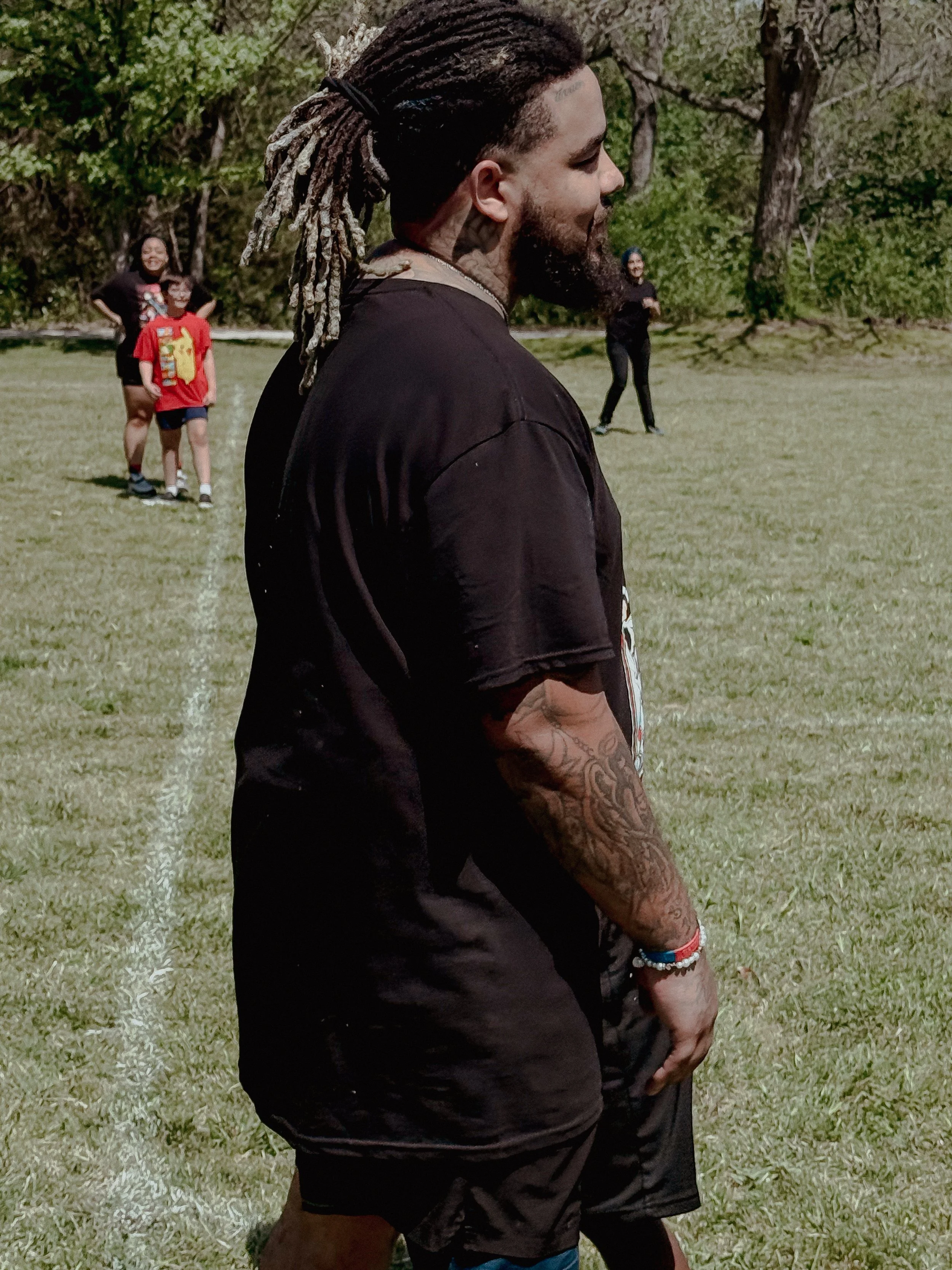 A man with dreadlocks, tattoos, and a beard standing on a grassy field, with three children and a woman in the background.