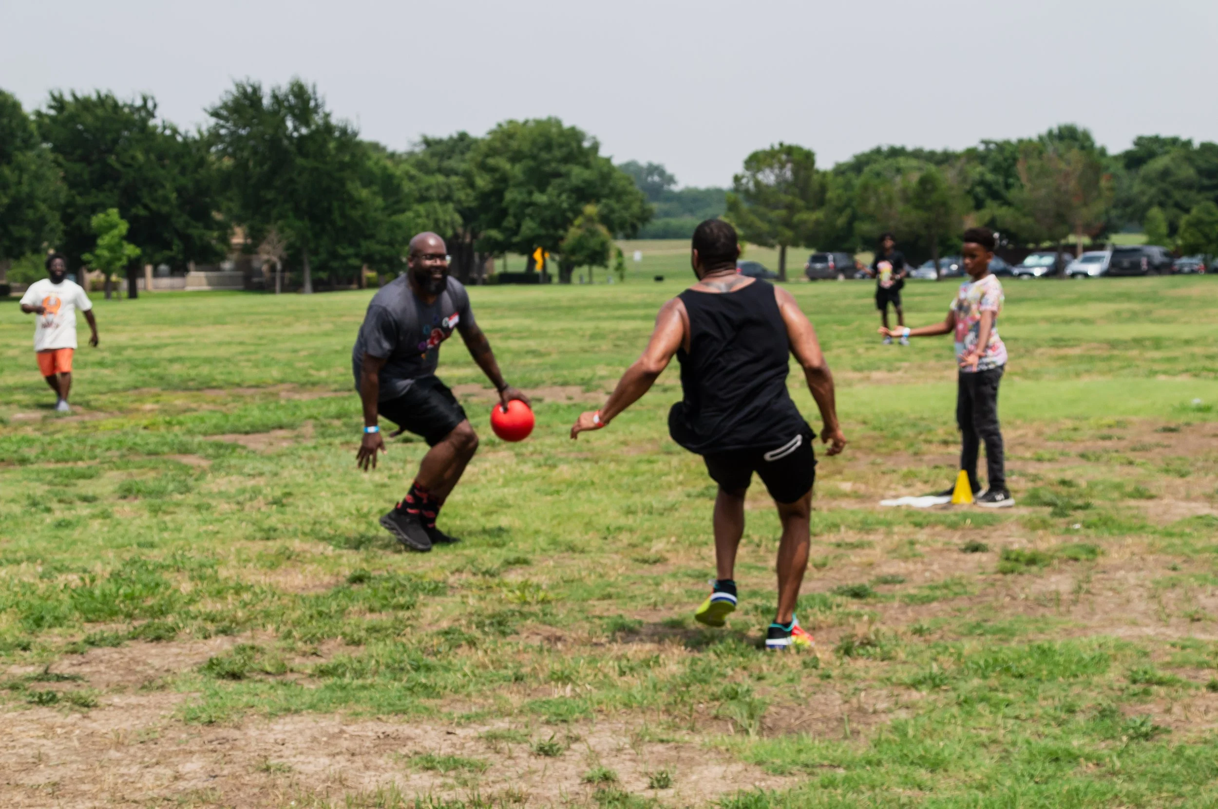 A group of people playing flag football outdoors on a grassy field, with trees and parked cars in the background.