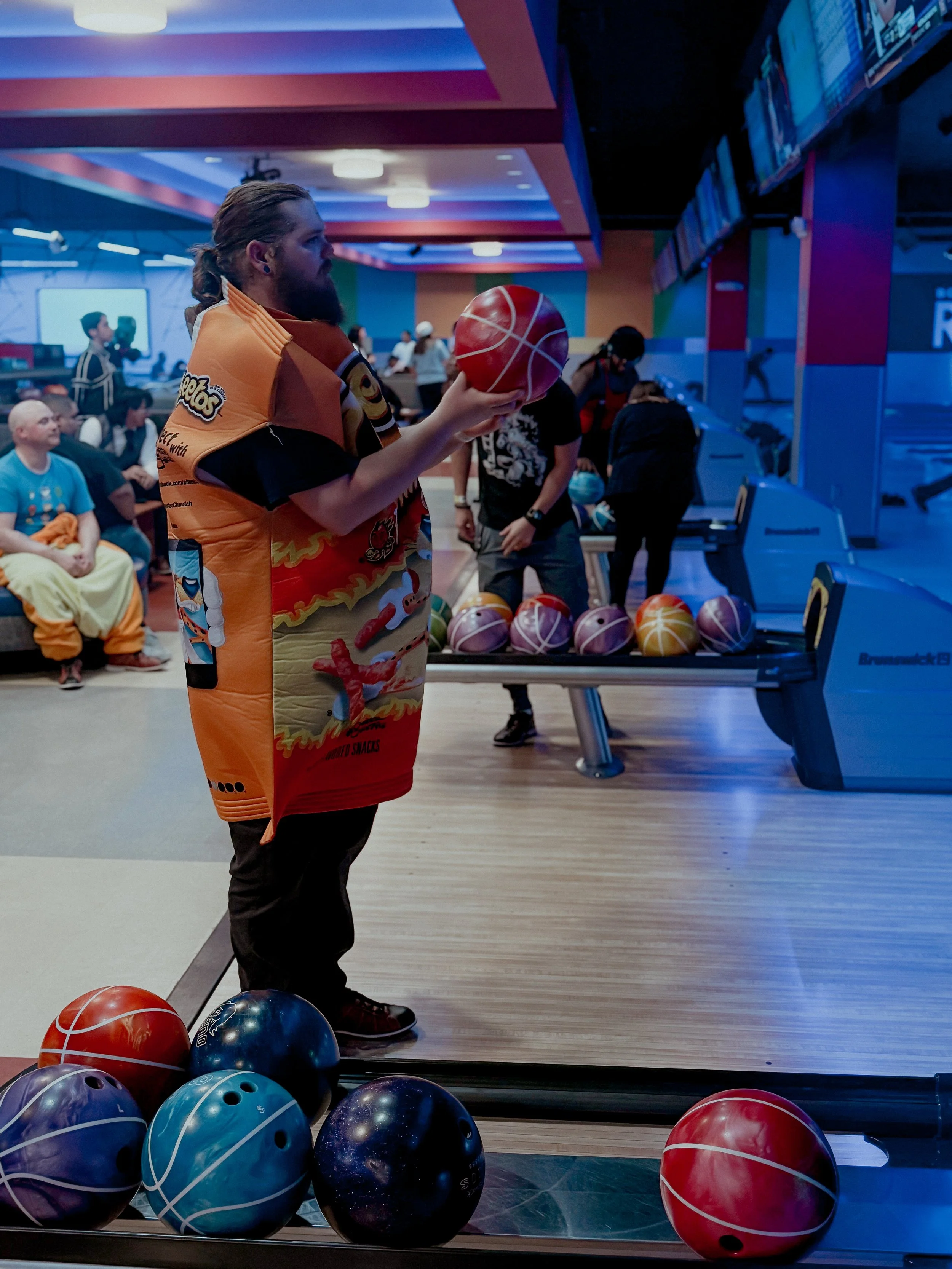A man with a beard and long hair in a ponytail is holding a bowling ball at a bowling alley, wearing a colorful bowling shirt with cartoon characters. Several bowling balls are on the ball return and on the floor nearby. In the background, other peop