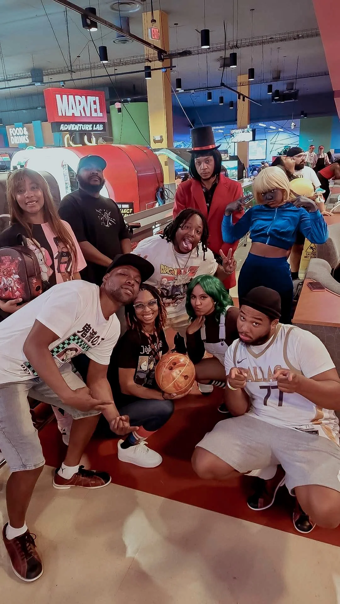 Group of people in cosplays and casual clothing posing together at an indoor arcade or entertainment center, with a Marvel Adventure Lab sign visible in the background.