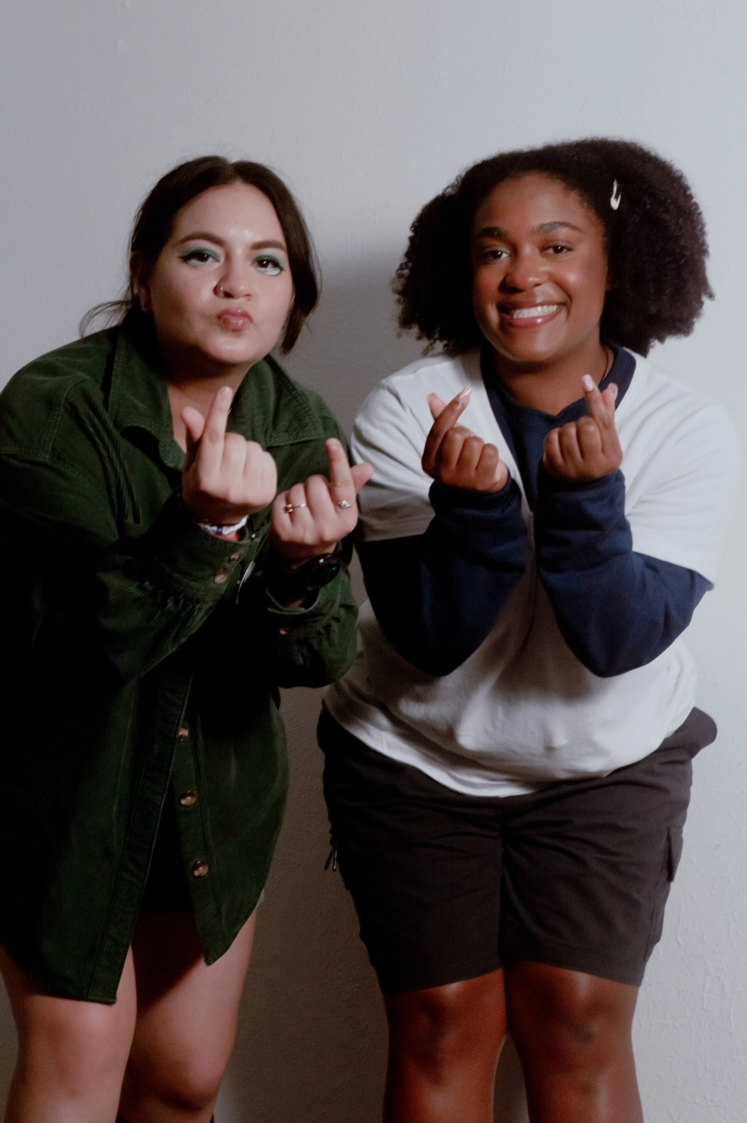 Two young women standing next to each other, making finger heart gestures, smiling, against a plain white wall.