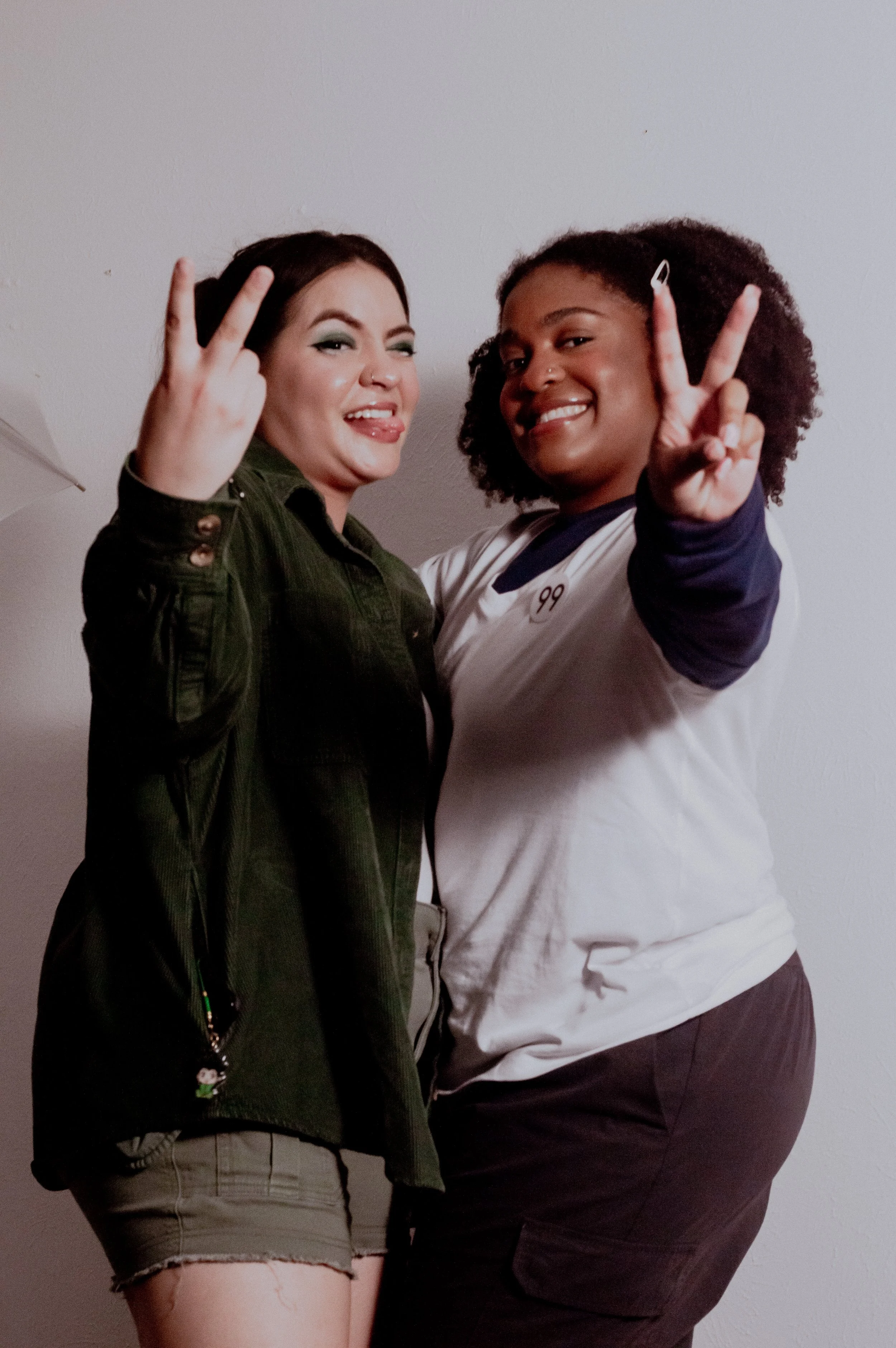 Two women standing close together, making peace signs with their hands, smiling, against a plain wall.
