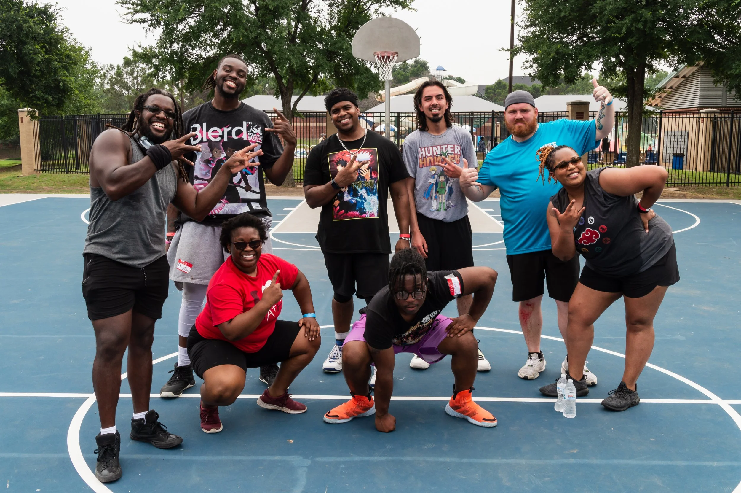 A group of nine people on an outdoor basketball court posing for a photo. They are smiling and making various hand gestures.