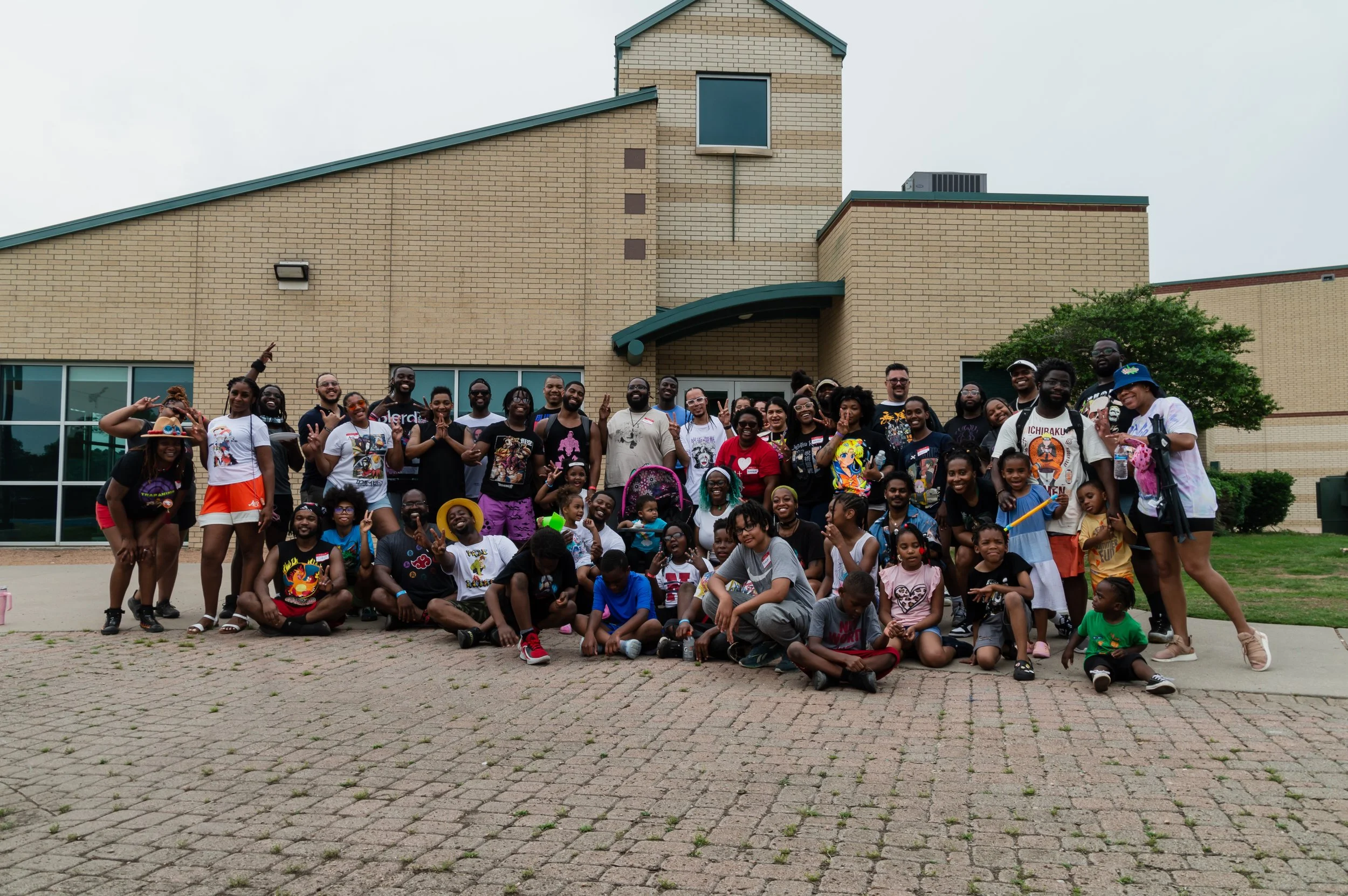 Group of children and adults posing for a photo outside a large brick building on a cloudy day.