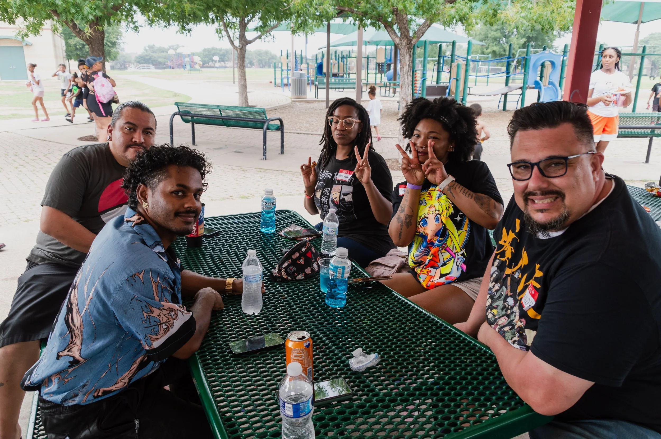 Group of five friends sitting at a green picnic table outdoors in a park, smiling and making peace signs, with playground equipment and children in the background.