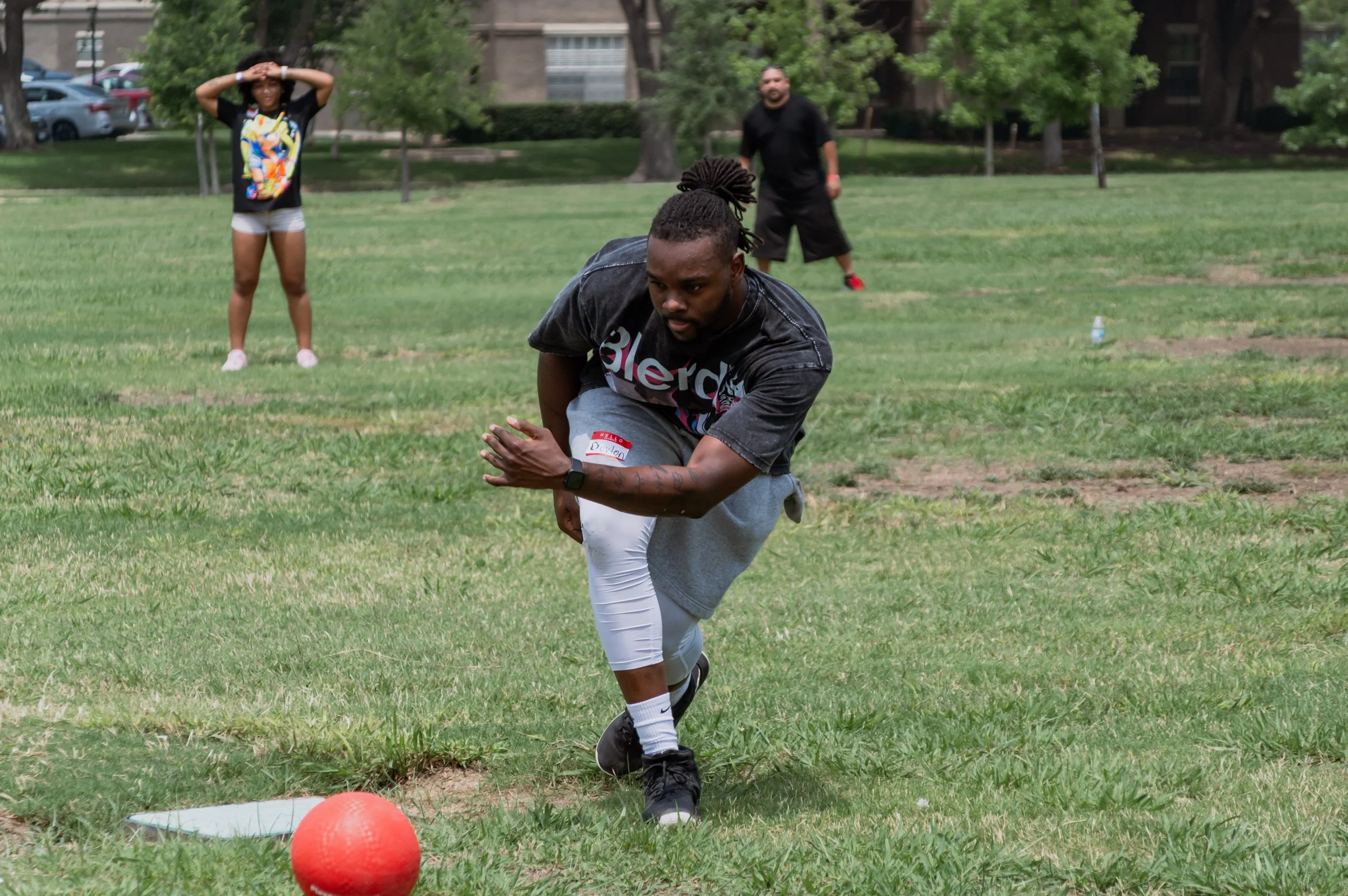 A man is stretching or running on a grassy field during an outdoor activity, with two other people in the background, a woman with her hands on her head and a man standing with his hands at his sides. The scene appears to take place in a park with tr