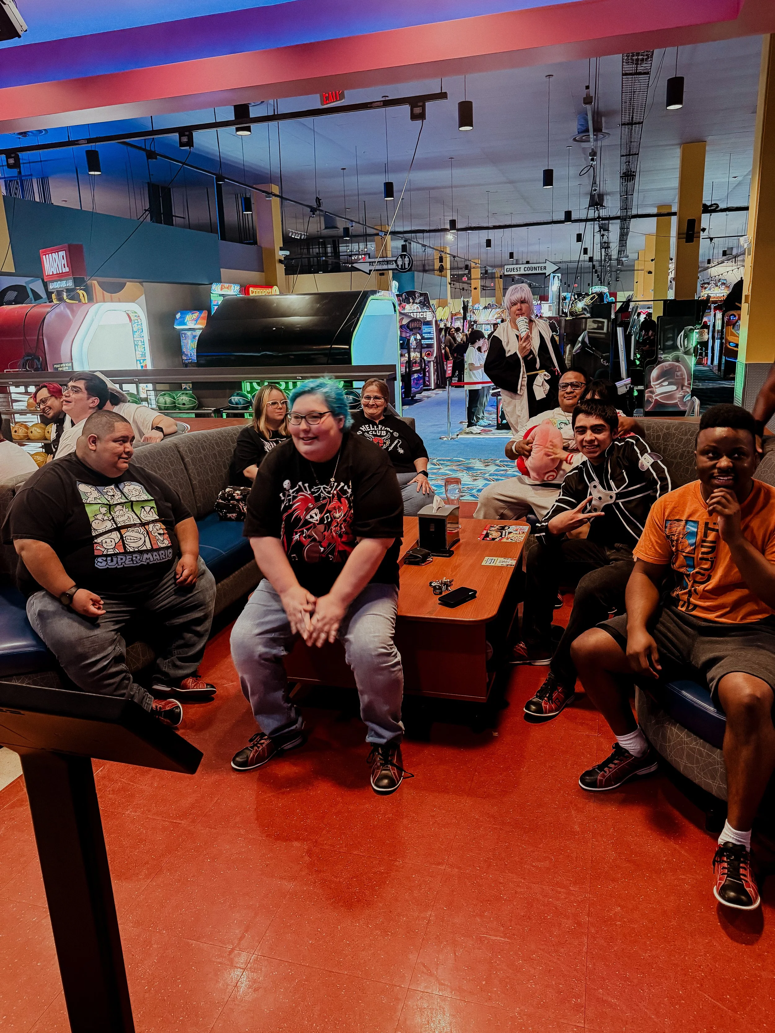 People sitting and standing around in a gaming arcade, with arcade machines and a woman singing karaoke.