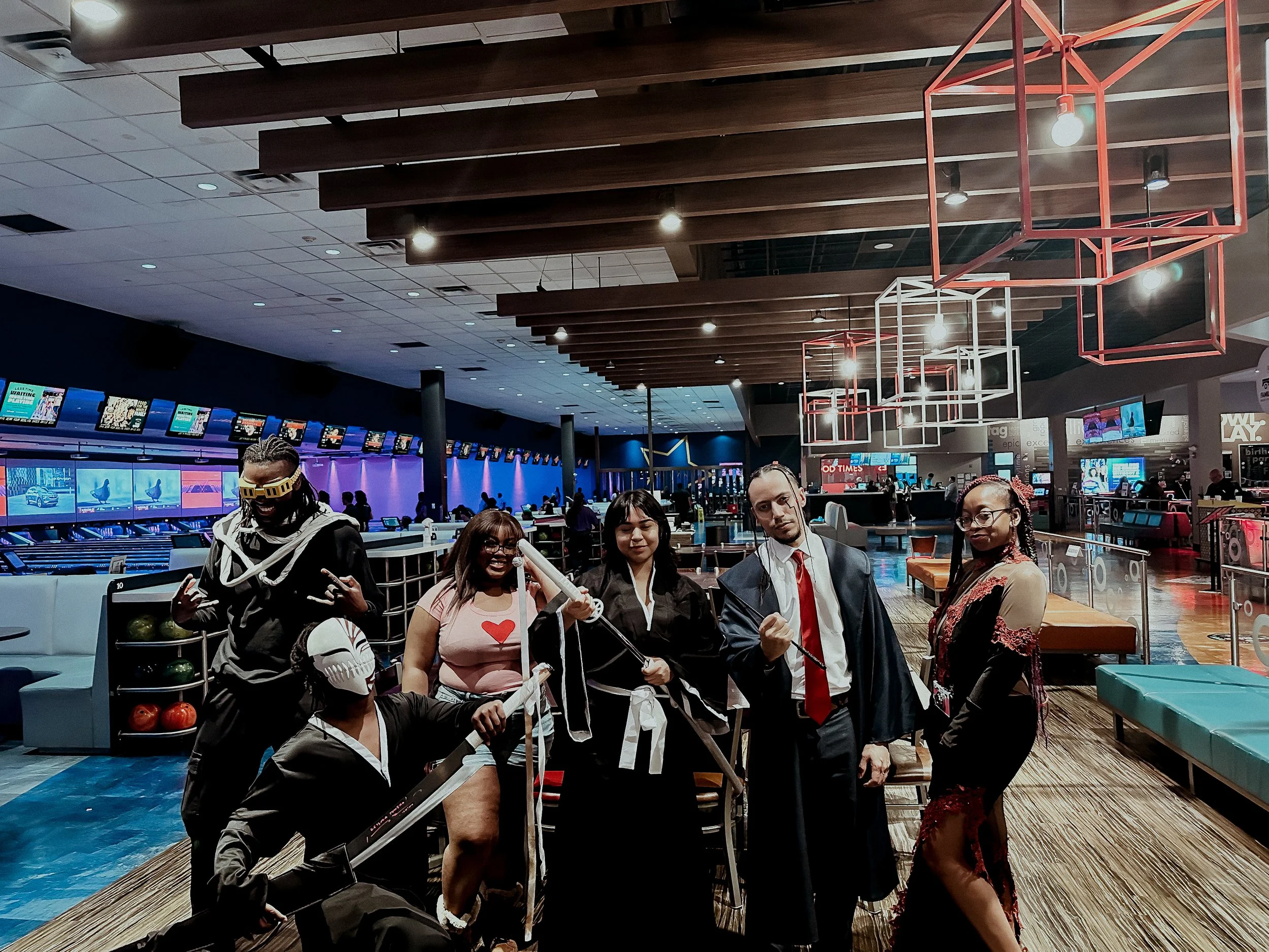 Group of people in costumes at a bowling alley, with bowling lanes and electronic scoreboards in the background.
