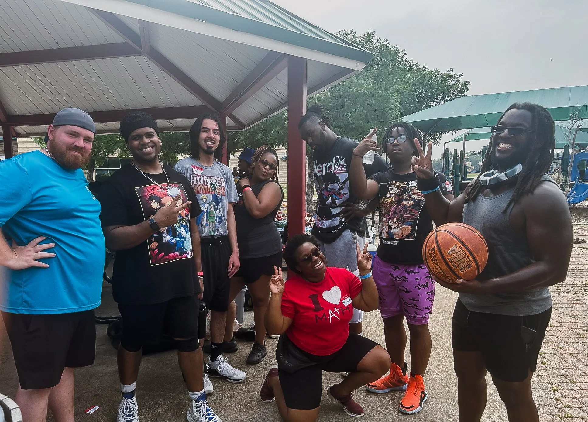 Group of diverse people at an outdoor basketball court, posing for a photo, with some making peace signs and smiling. One person is holding a basketball. There is a playground in the background.