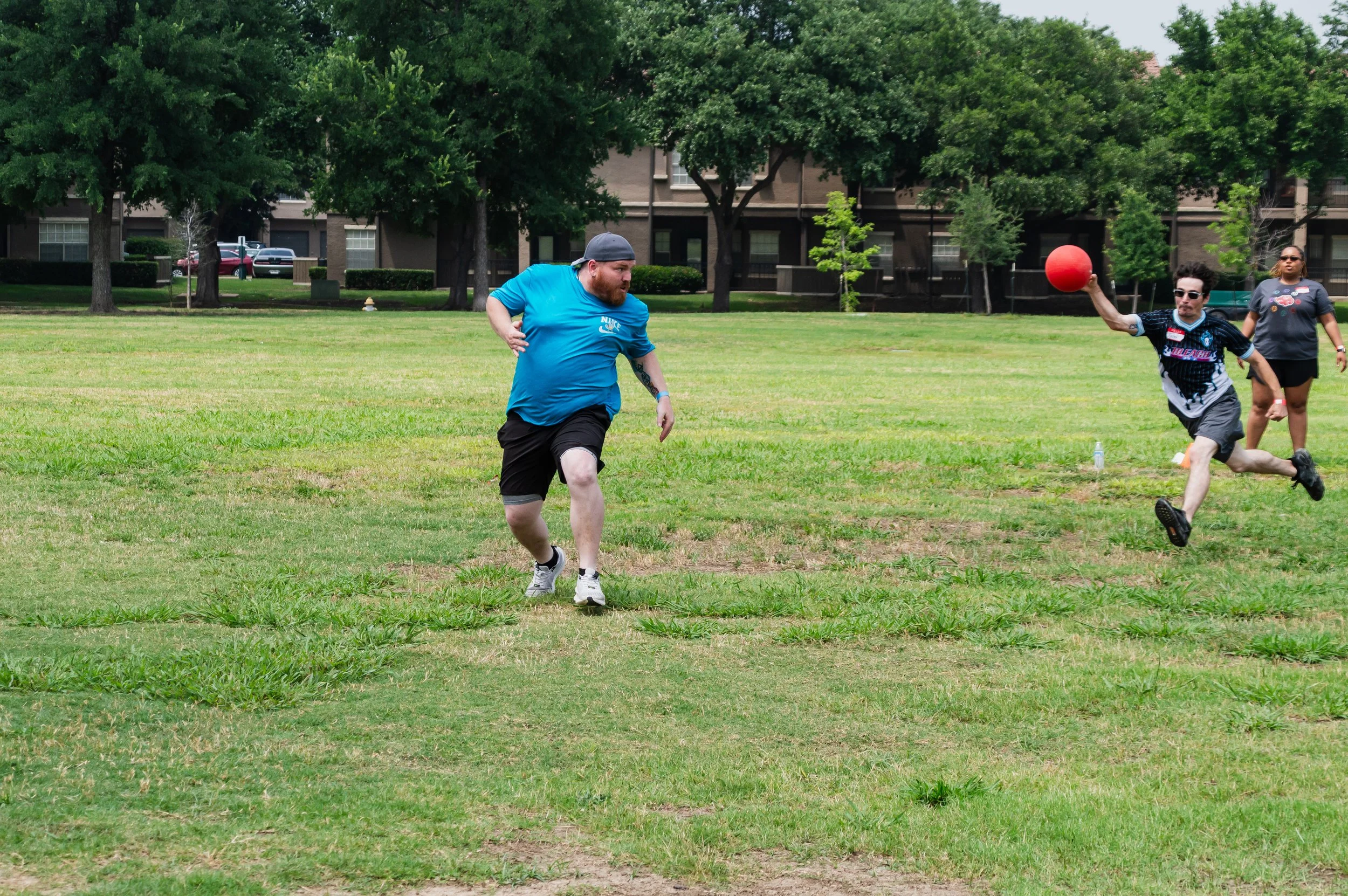 People playing kickball on a grassy field with trees and apartment buildings in the background.