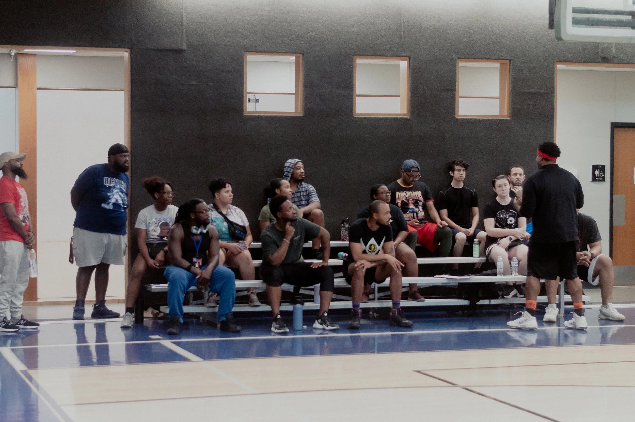 Group of people sitting on bleachers inside a gymnasium, listening to a person standing and speaking in front of them.