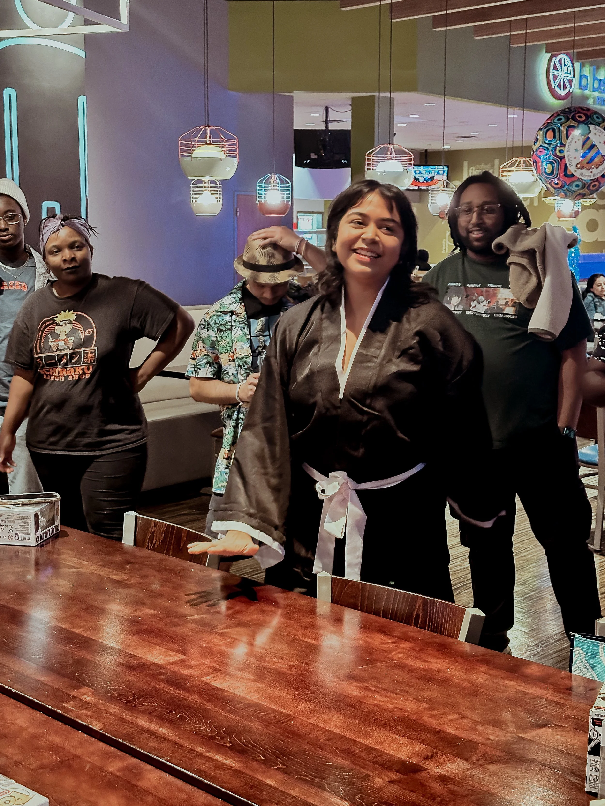 A woman in a black robe with a white ribbon tied around her waist, smiling and gesturing with her hand, standing at a table in a restaurant or café with several other people in the background.