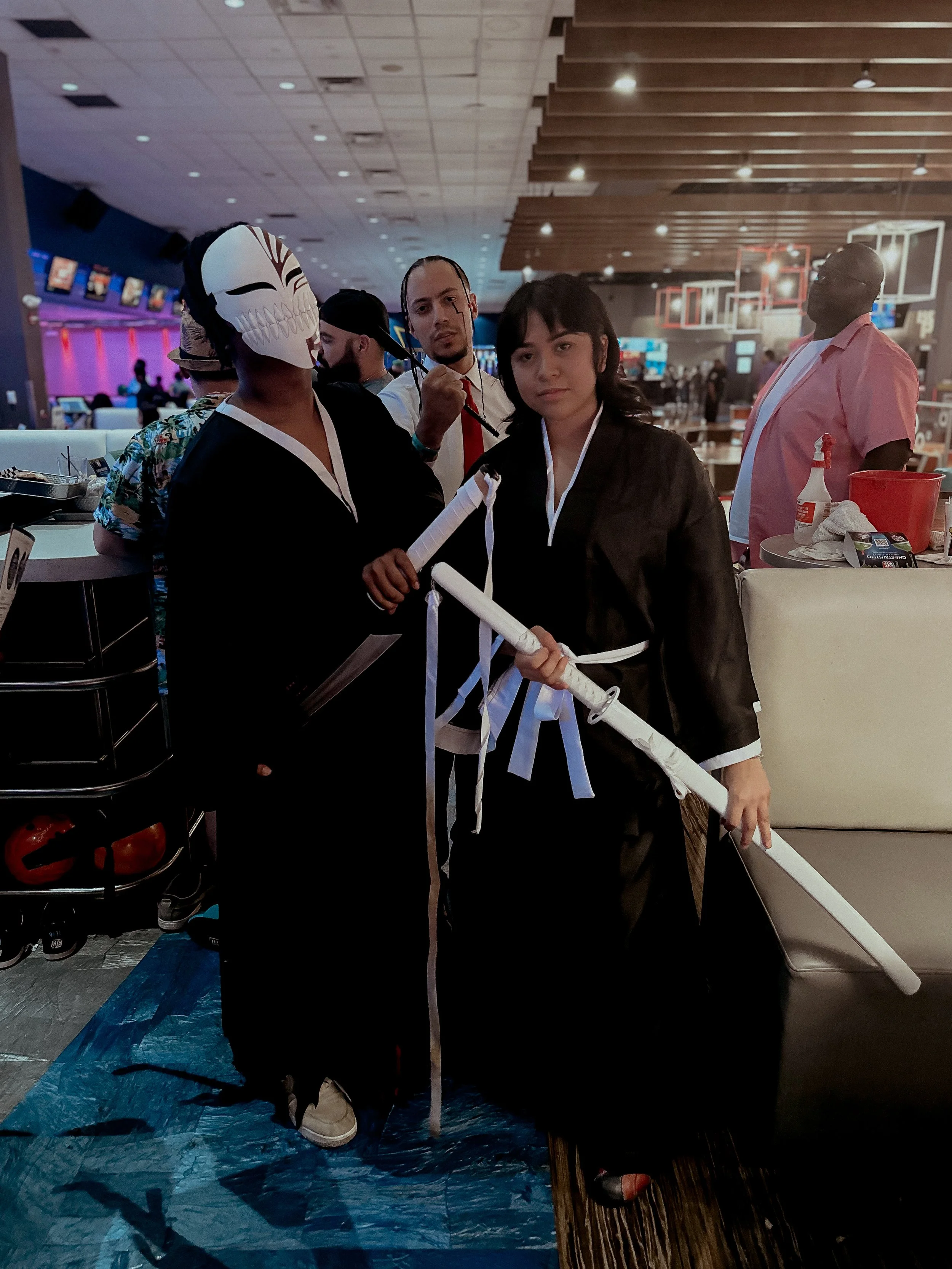 Group of people in costume at a bowling alley, with one person wearing an Oni mask and holding a katana, and others in cosplay attire.