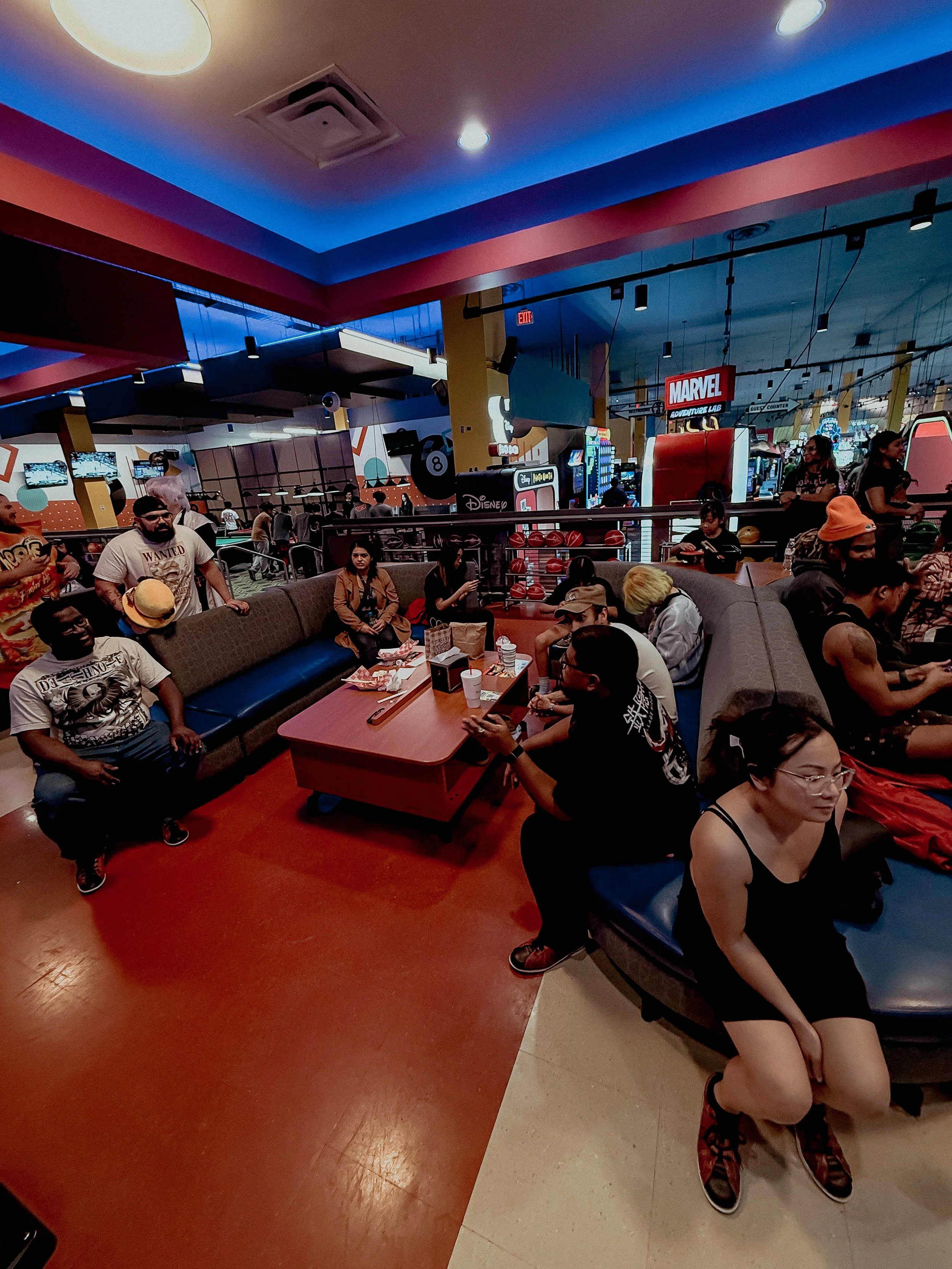 People sitting and standing in an arcade lounge area with gaming stations and colorful signage in the background.