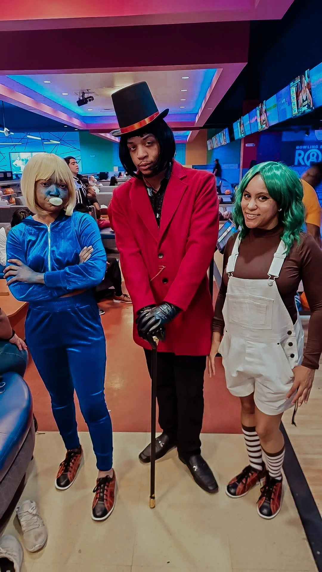 Three people dressed in cosplay costumes, one person in a clown costume with blonde wig and blue face paint, one person as a magician with a top hat and red coat, and one person with green wig and striped socks, at a bowling alley.