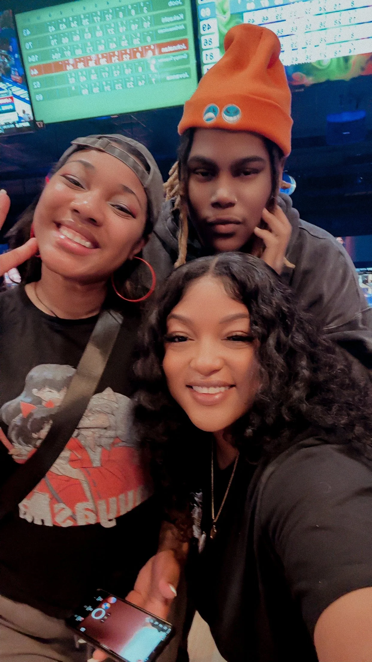 Three people smiling and posing for a selfie in front of a colorful electronic scoreboard, with one person making a peace sign.