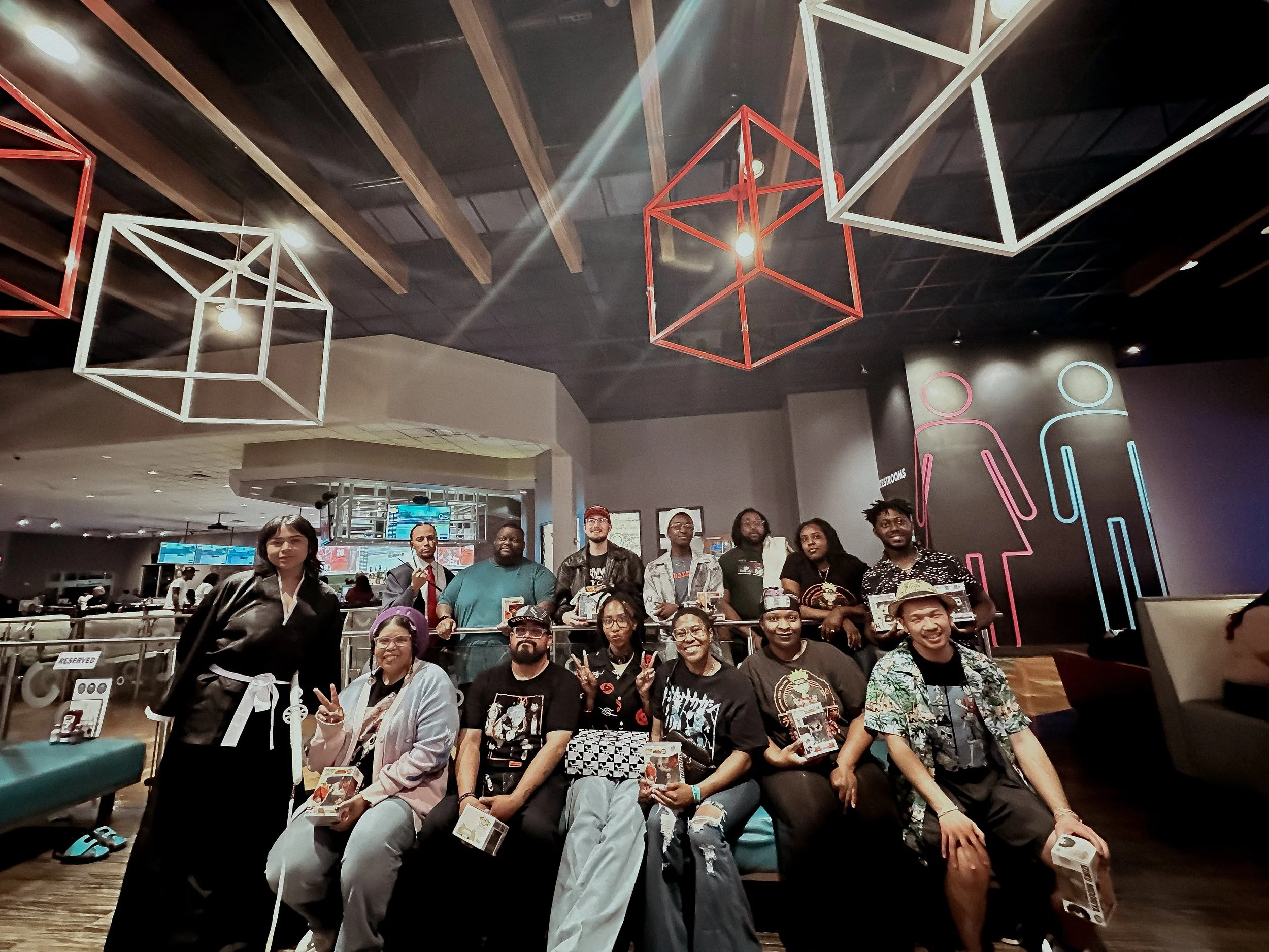 Group of people posing for a photo in a modern indoor space, some sitting and some standing, with a neon sign of restroom symbols on the wall behind them and geometric ceiling lights overhead.