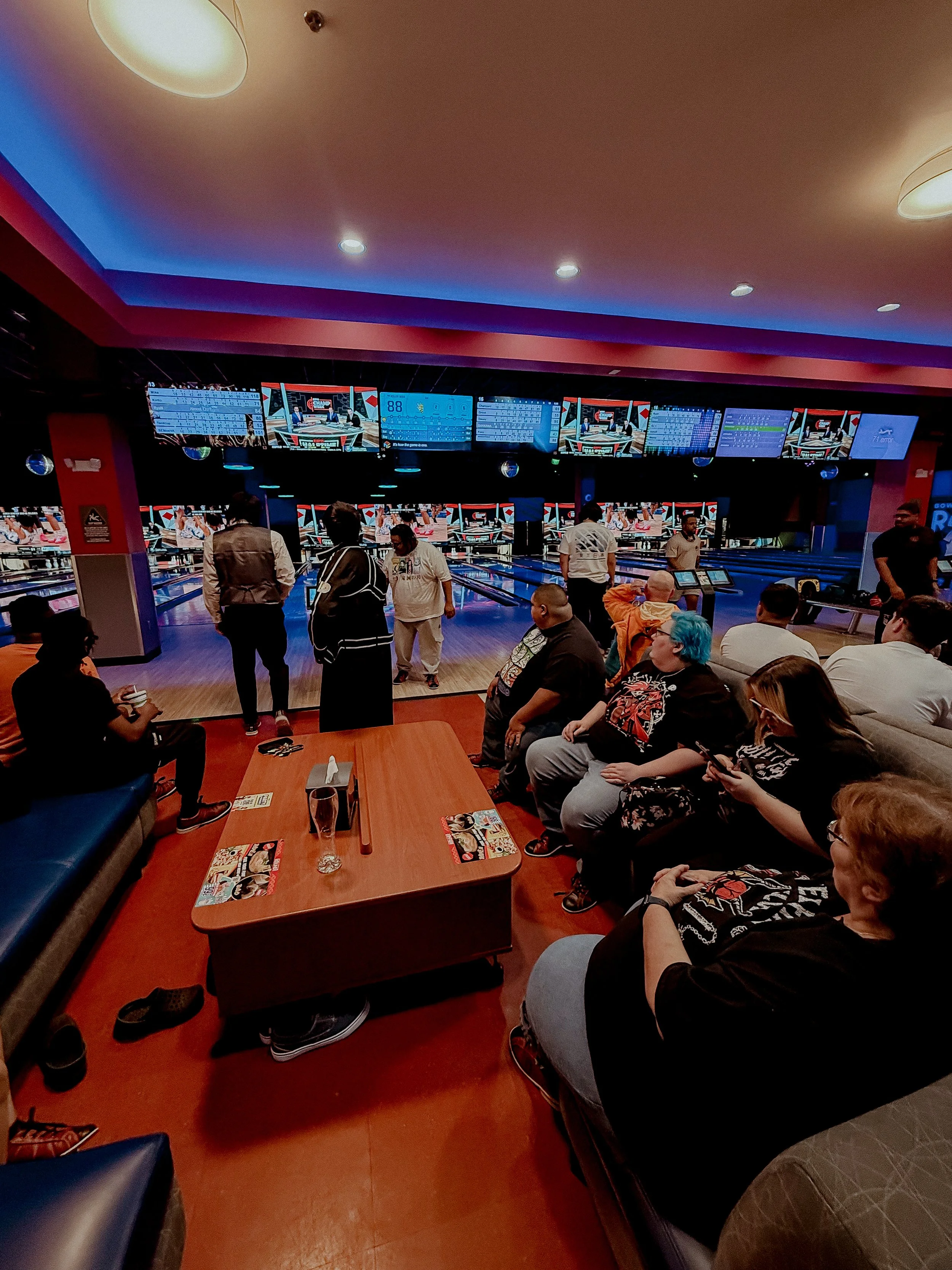 People at a bowling alley, some sitting on couches and chairs watching others bowl, with multiple monitor screens displaying scores and game information in the background.