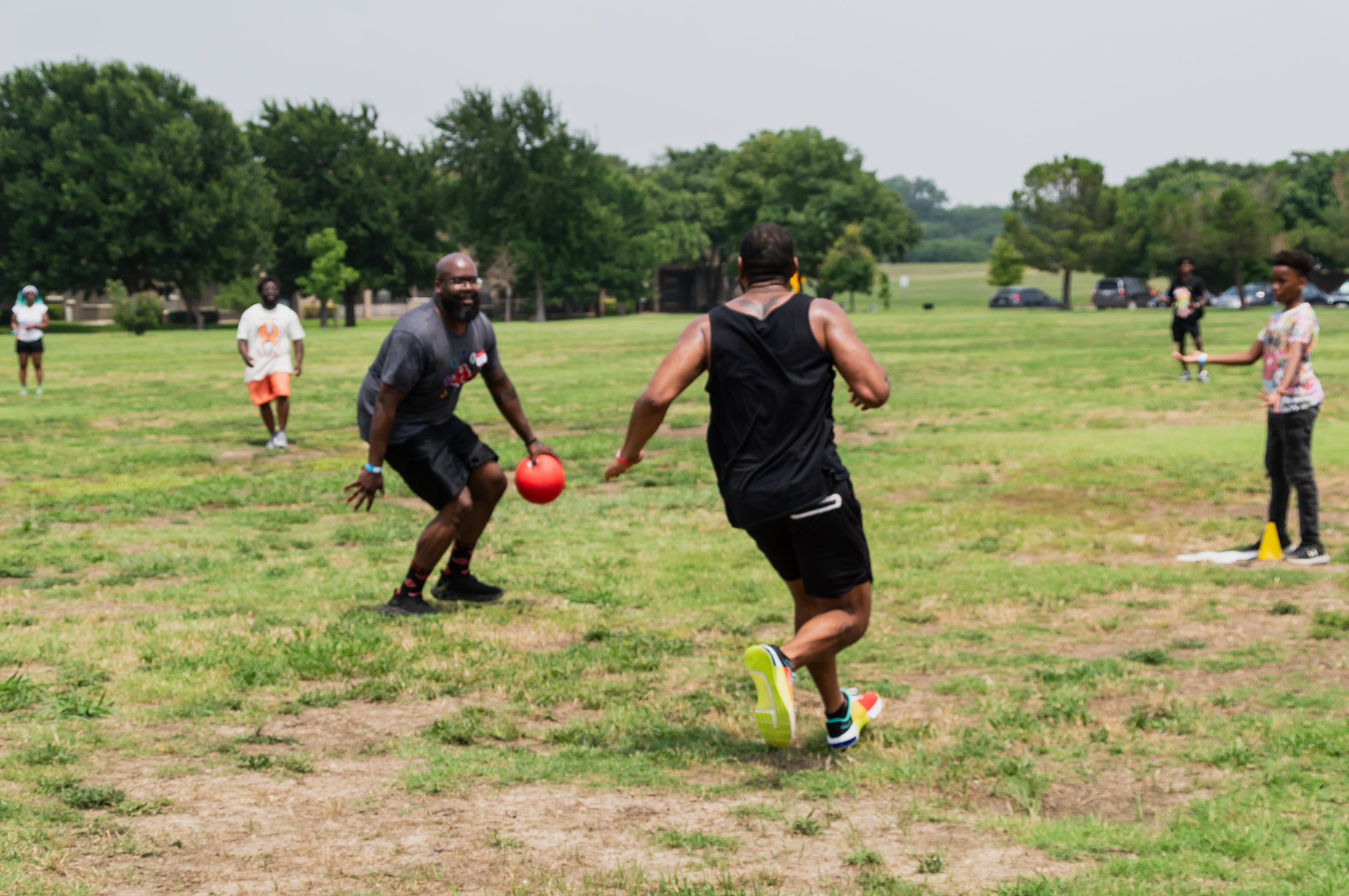 Group of people playing kickball on a grassy field in a park with trees and cars in the background.