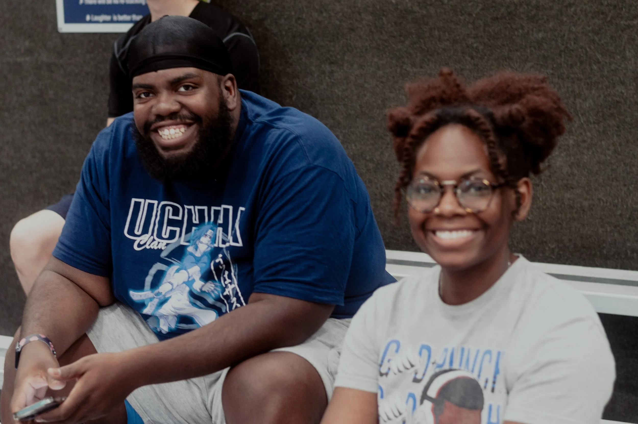 Two young adults sitting on a bench at an indoor location, smiling at the camera. The man wears a blue T-shirt and a black durag, while the woman wears glasses and a light-colored T-shirt with a graphic.