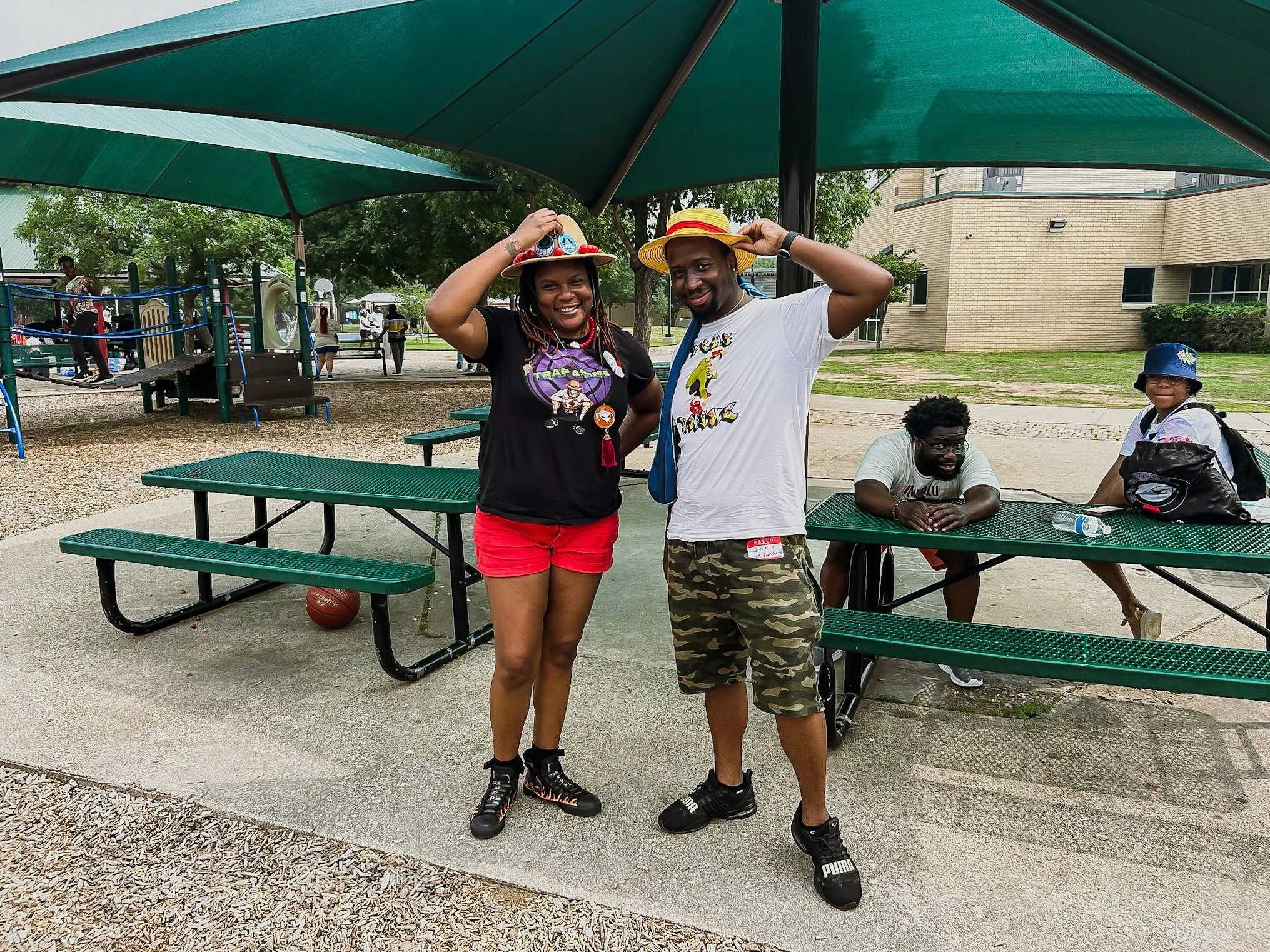 Two people smiling and posing for a photo under a large green canopy at a park, wearing hats. Two other individuals are sitting at a green park bench nearby, one leaning on the table and the other standing with a backpack.