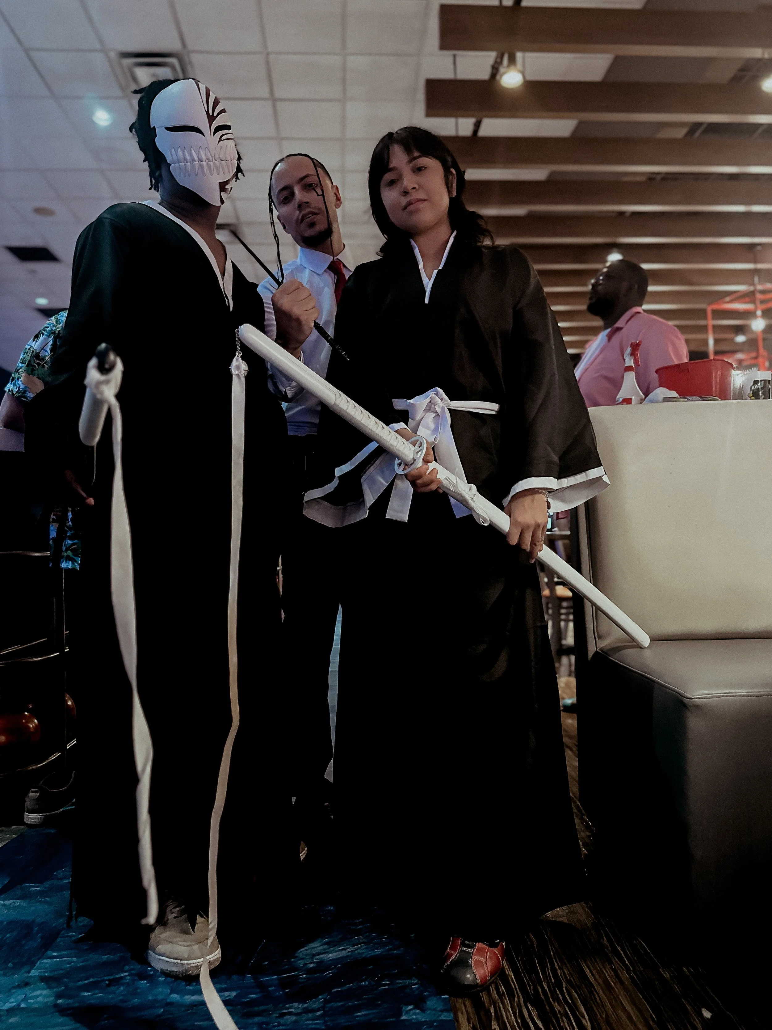 Three people in costumes posing together at a gathering. The person on the left wears a skull mask and black outfit, holding a weapon. The person in the middle has dreadlocks, a white shirt, and red tie. The person on the right wears a black robe wit