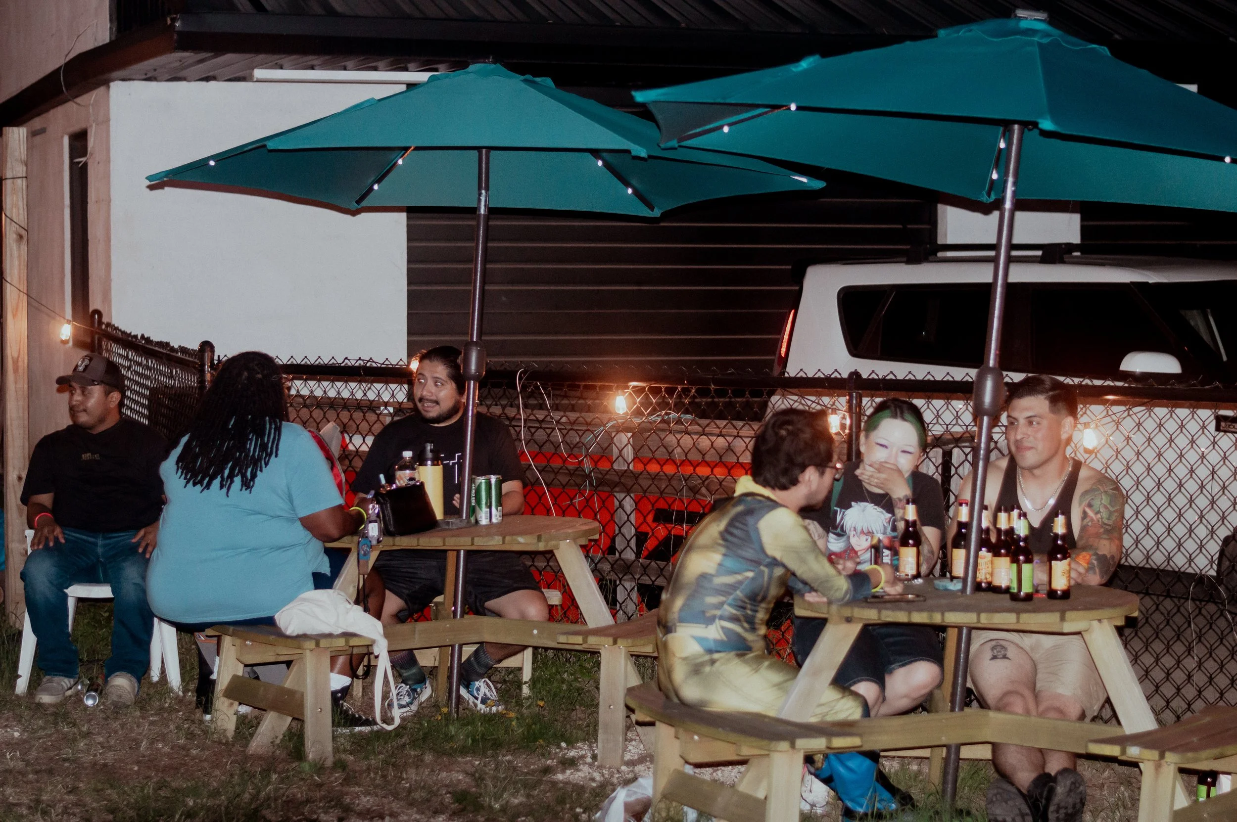 Group of friends sitting at picnic tables under teal umbrellas, socializing and drinking outdoors at night.