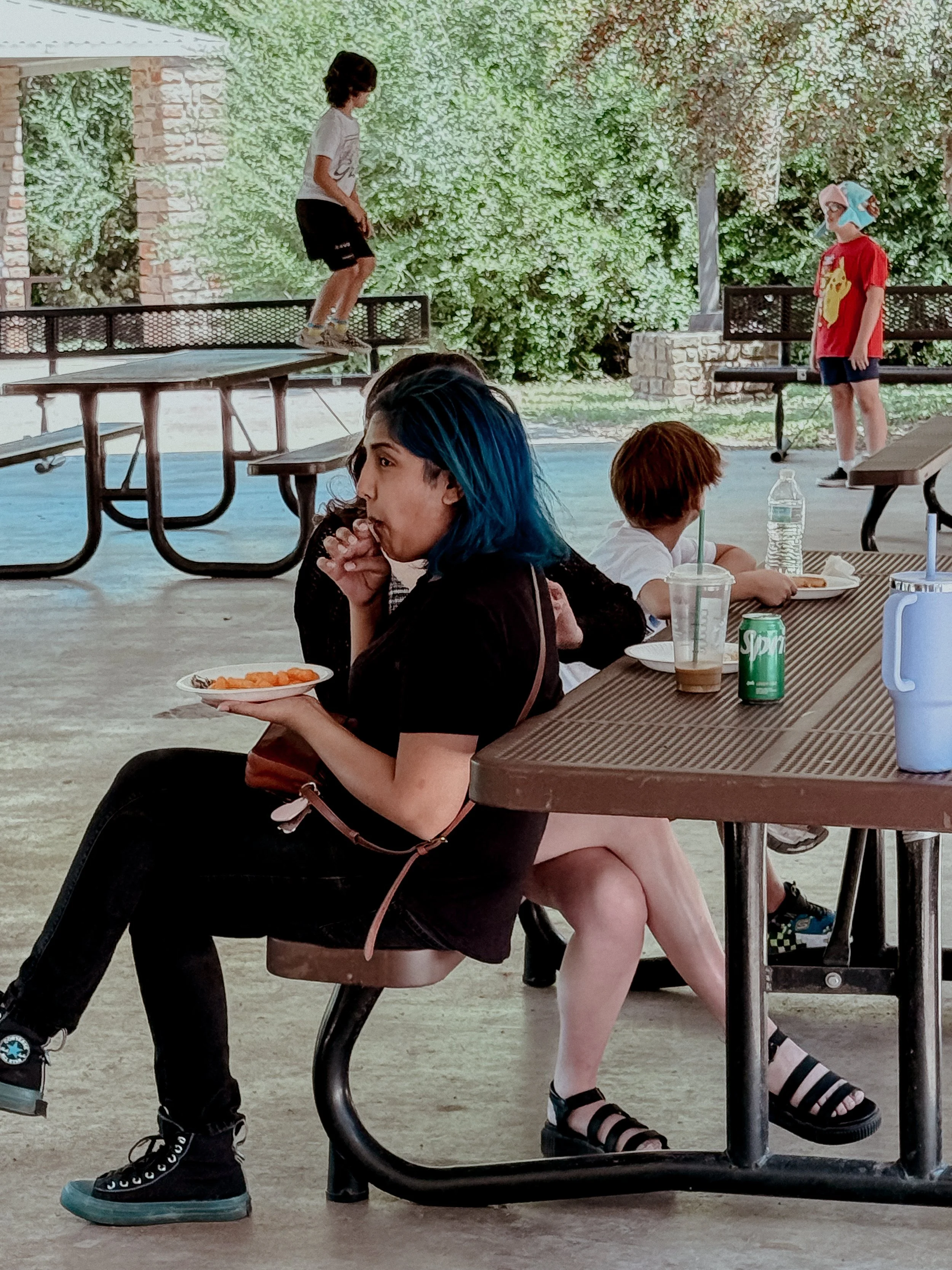 A woman with blue hair sitting at a picnic table eating snack, children in the background, one on a skateboard on a table and another wearing a hat and standing outside near trees.