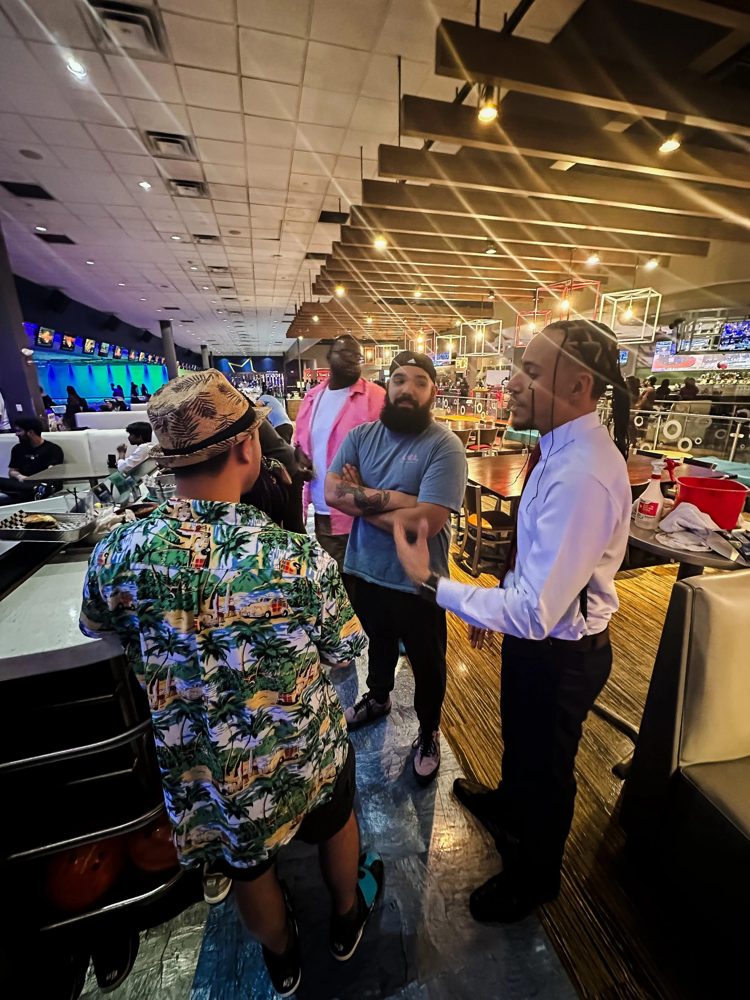 Four men are standing and talking in a brightly lit bowling alley, with bowling balls and lanes in the background. One man is dressed in a white shirt and has long hair tied back, while another is wearing a casual gray T-shirt and a black backwards c