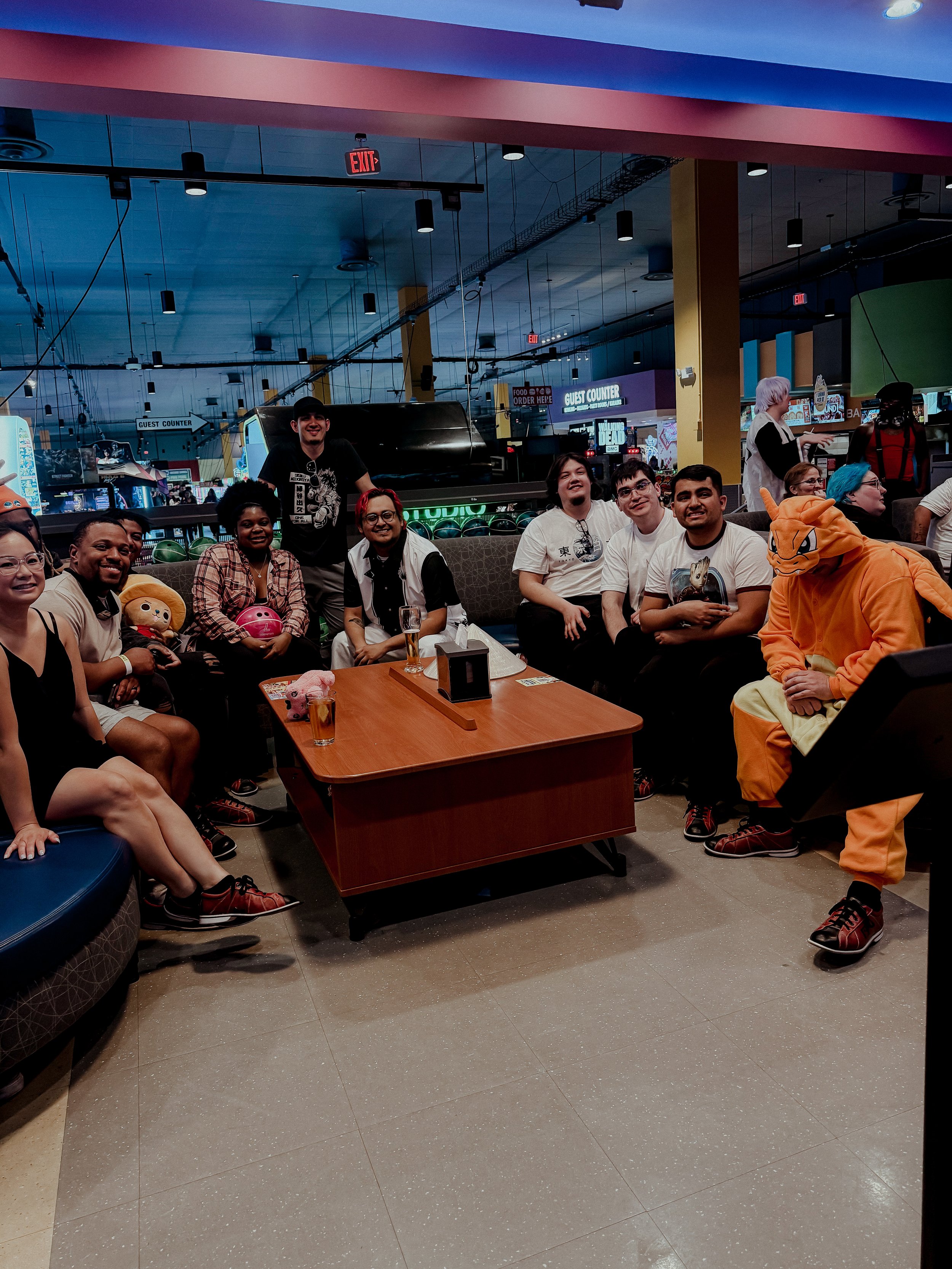 Group of people, some in costumes, sitting and standing around a table in an arcade or entertainment venue.