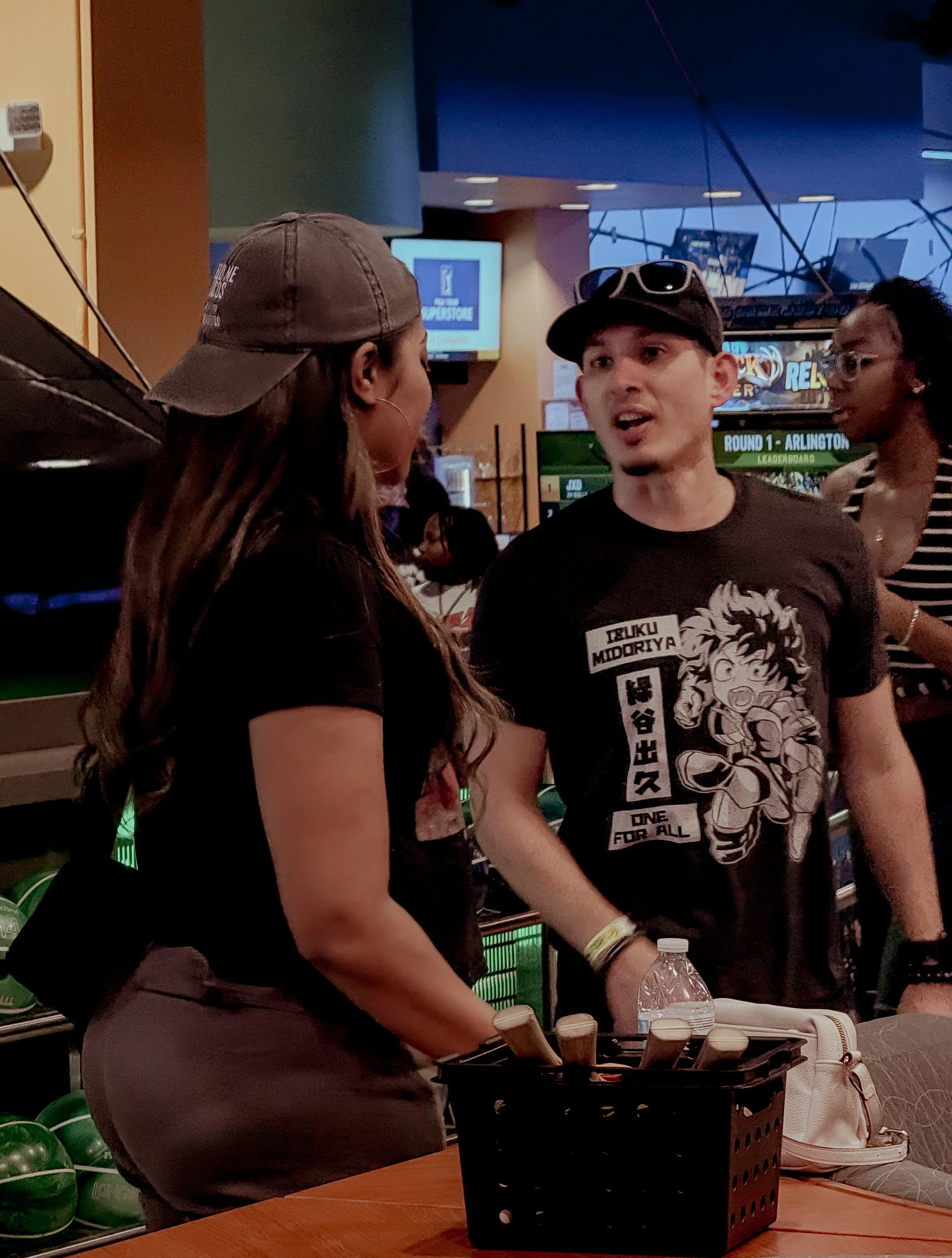 A man and woman having a conversation at a store, with the woman wearing a baseball cap and the man wearing sunglasses on his cap. There are watermelon and shopping items on the table in front of them, and other people and screens in the background.